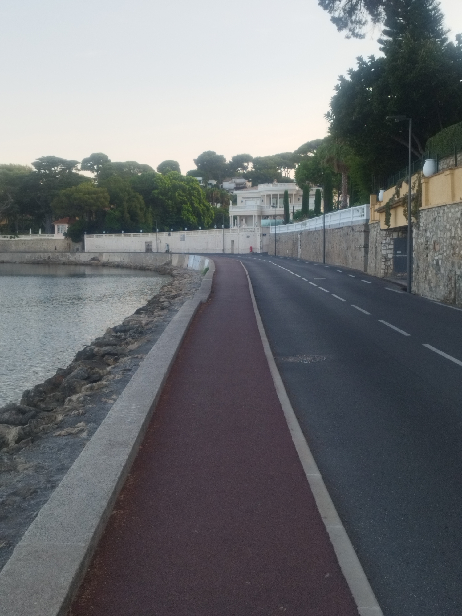 The image depicts a coastal road and a pedestrian path in Antibes, France, with the Mediterranean Sea on the left and a street with a red-colored pedestrian lane on the right. The sea is calm with gentle ripples. A stone-lined embankment separates the sea from a concrete walkway. Beyond the walkway, a white wall runs along the edge of the property, with lush green trees and elegant white buildings visible behind it. The road is paved with asphalt and has white dashed lane markings. A stone wall topped with greenery borders the right side of the road, and further back, a yellow building with stone accents can be seen. The sky is a pale, overcast hue, suggesting early morning or late afternoon light. No people or vehicles are present in the scene.