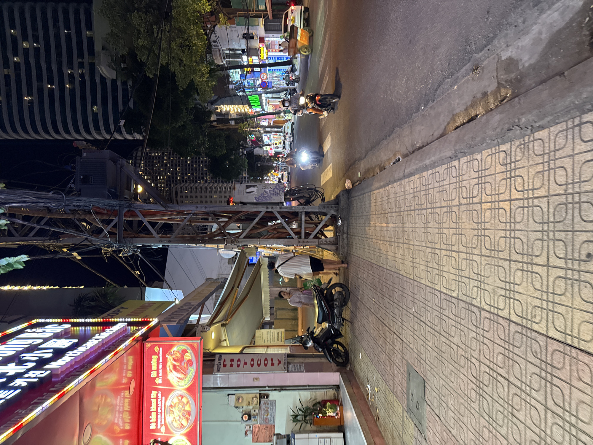 The image depicts a street scene at night in Nha Trang, Vietnam, likely on a commercial street given the illuminated signs and buildings. A pedestrian sidewalk with patterned tiles is in the foreground, with a street running alongside it. On the sidewalk, a black motorcycle is parked. Two individuals are walking away from the camera on the sidewalk, one wearing a striped shirt and shorts, and the other in lighter clothing. To the left, a brightly lit sign for "NORTHEAST CHICKEN" is visible, featuring images of food. Adjacent to this is a building with a "PHOTOCOPY" sign.
The street is moderately busy with traffic. Several motorcycles are visible, some with riders. One motorcycle, in the middle ground, is heading towards the camera, with its headlight illuminated. Another motorcycle is further down the street, also with its headlight on. Cars and other vehicles are present in the background, along with more illuminated signs, indicating commercial establishments. Tall buildings line the street in the distance, some with visible windows lit up. Electrical wires and poles are prominent in the mid-ground, contributing to the urban atmosphere. The overall lighting suggests it is after dusk, with artificial lights illuminating the scene.