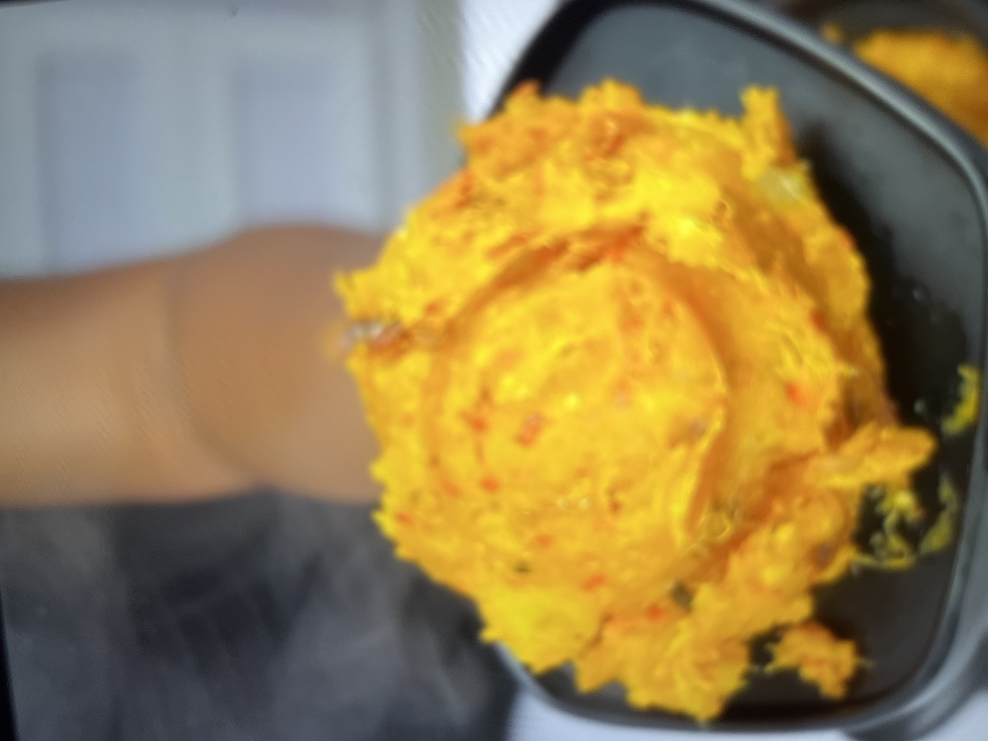 The image shows a close-up of a brightly colored, fluffy yellow substance with small red flecks, placed inside a dark, rectangular container. The substance appears to be a food item, possibly mashed or whipped. There is a hint of steam rising from the food, suggesting it is hot. In the background, out of focus, is a blurred beige and light blue area that could be a wall or a soft surface. To the left of the frame, a blurred section of what appears to be an arm or hand, rendered in a warm, brownish tone, is visible. The lighting in the image is diffused, creating soft shadows. The overall impression is of a food preparation or serving scene. No text is visible in the image. The provided location context is Abuja, Nigeria.