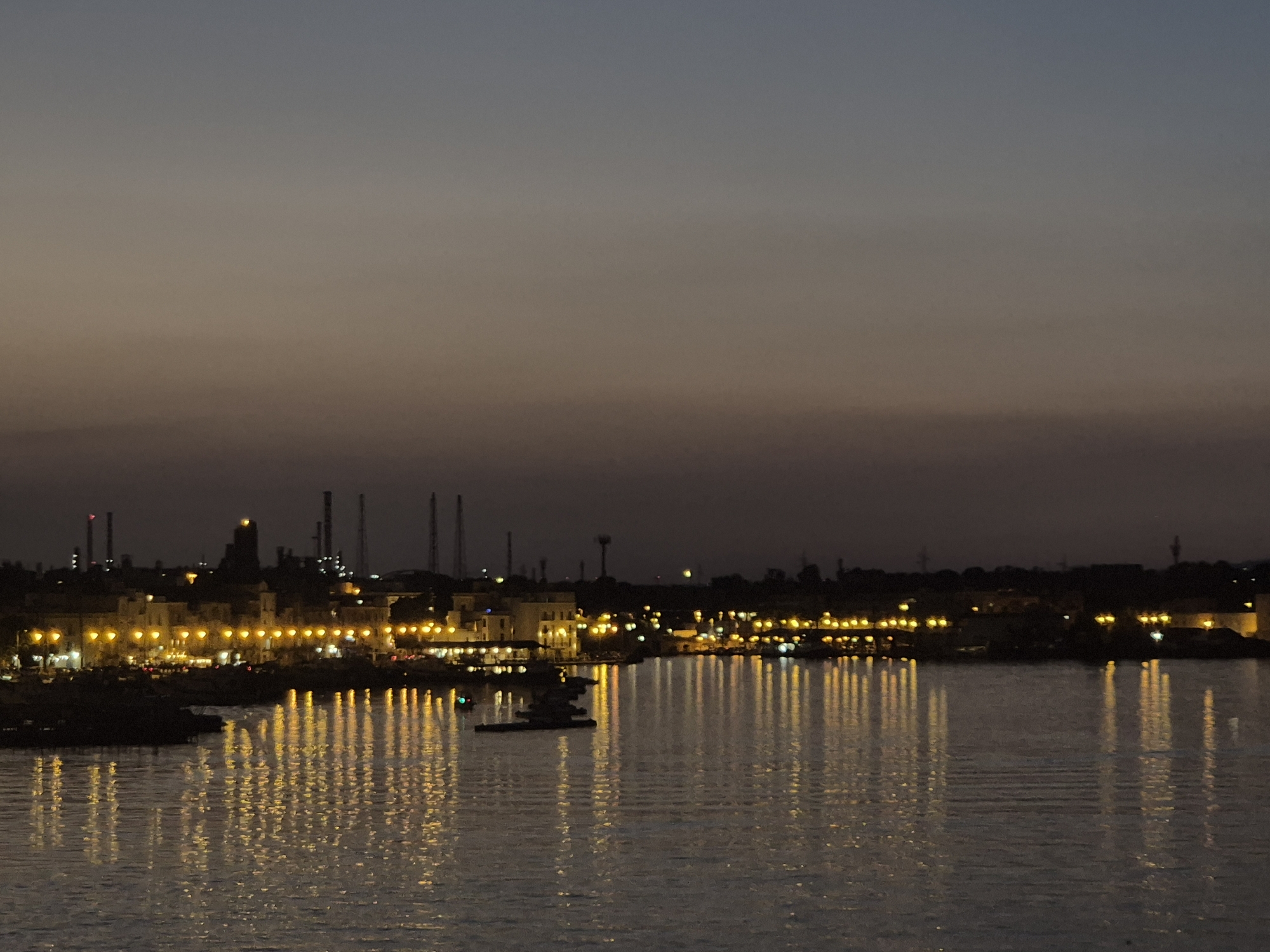 The image captures a twilight view of the waterfront in Taranto, Italy, likely in the Mar Piccolo area given the presence of industrial structures. The sky transitions from a deep blue at the top to a hazy, lighter gradient of grey and tan near the horizon, indicating the last vestiges of daylight. The water in the foreground reflects the numerous yellow lights from the buildings and streetlamps lining the distant shore.
The shoreline is densely populated with buildings of varying heights and architectural styles, many of which are illuminated with warm yellow lights. Beyond the buildings, several tall industrial structures, resembling smokestacks and towers, punctuate the skyline, silhouetted against the fading sky. The reflections of the lights on the water create a shimmering, almost abstract pattern of vertical streaks.
In the middle of the water, a dark, low-lying structure, possibly a barge or a floating platform, is visible. A small boat with a green light is also present in the water, further to the left. The overall impression is one of a coastal city at dusk, with the industrial backdrop contrasting with the illuminated urban waterfront.