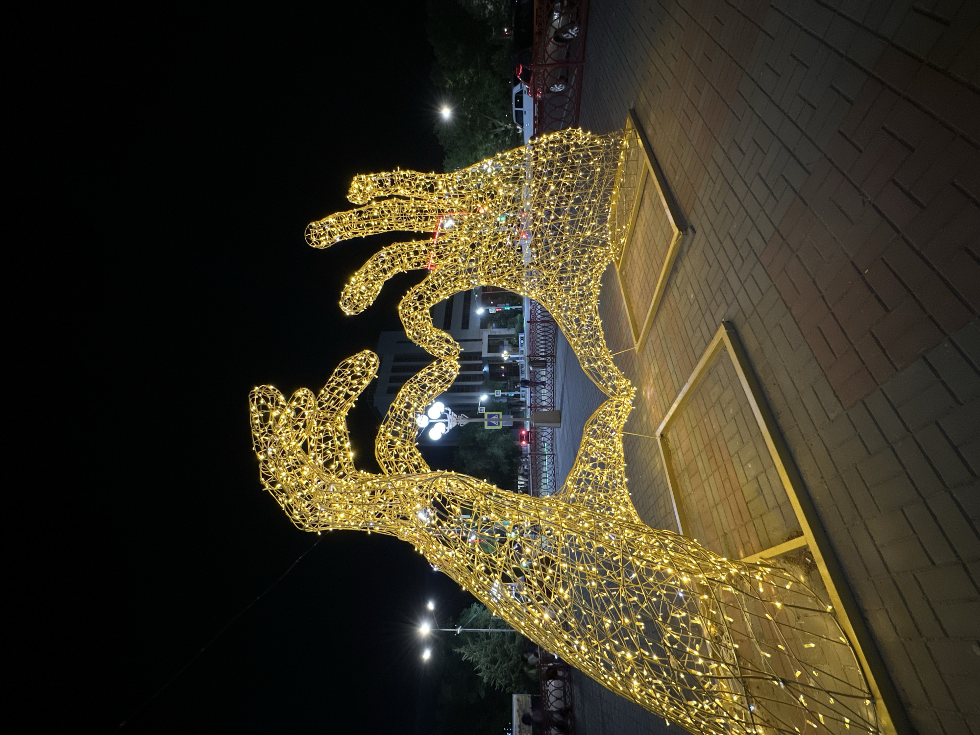 An illuminated sculpture of two hands forming a heart shape stands in a city plaza at night. The sculpture is made of wire mesh and is covered in warm yellow LED lights, creating a glowing effect. The hands are large and stylized, with fingers slightly bent to form the heart. The sculpture is supported by a golden metal frame on a paved surface. In the background, streetlights illuminate the area, and trees and buildings are visible. A few cars and pedestrians are present in the distance. The overall scene suggests a festive or decorative public space, likely in a city. This sculpture is located on Arbat Street in Elista, Russia, a pedestrian zone known for its art installations and public spaces.