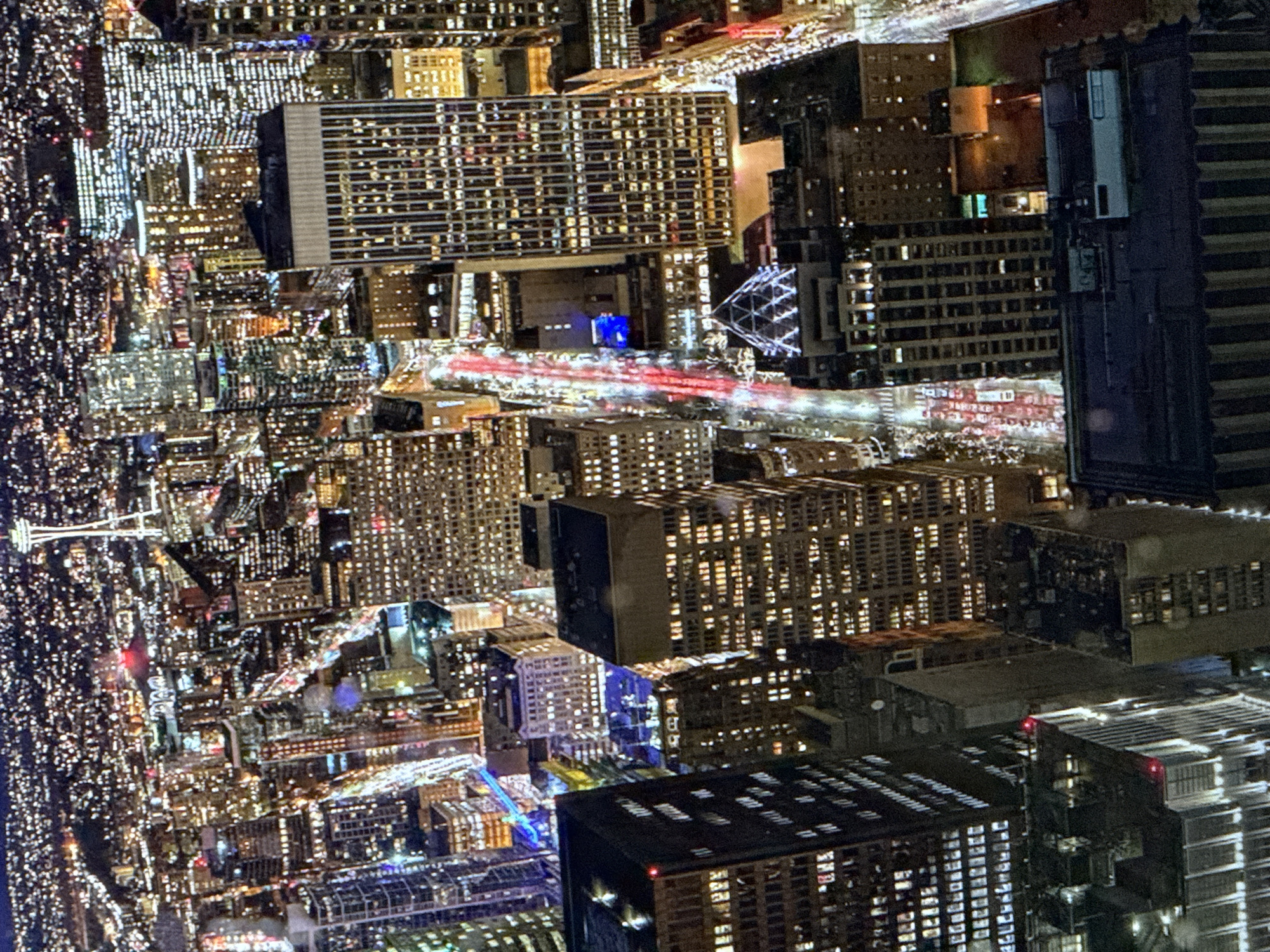 The image is an aerial, nighttime view of a dense urban cityscape, likely a major city. The scene is dominated by numerous skyscrapers illuminated by countless windows, creating a vibrant tapestry of light against the dark sky. A prominent landmark resembling the Space Needle in Seattle, Washington, is visible in the distance, topped with a glowing light. Streets below are illuminated by the lights of buildings and the red and white streaks of vehicle headlights and taillights, indicating ongoing traffic. Various architectural styles are present, from modern glass structures to older, more ornate buildings. The overall impression is one of a bustling metropolis at night. The user has requested the location to be Saratov, Russia, but the visual cues strongly suggest Seattle, Washington.