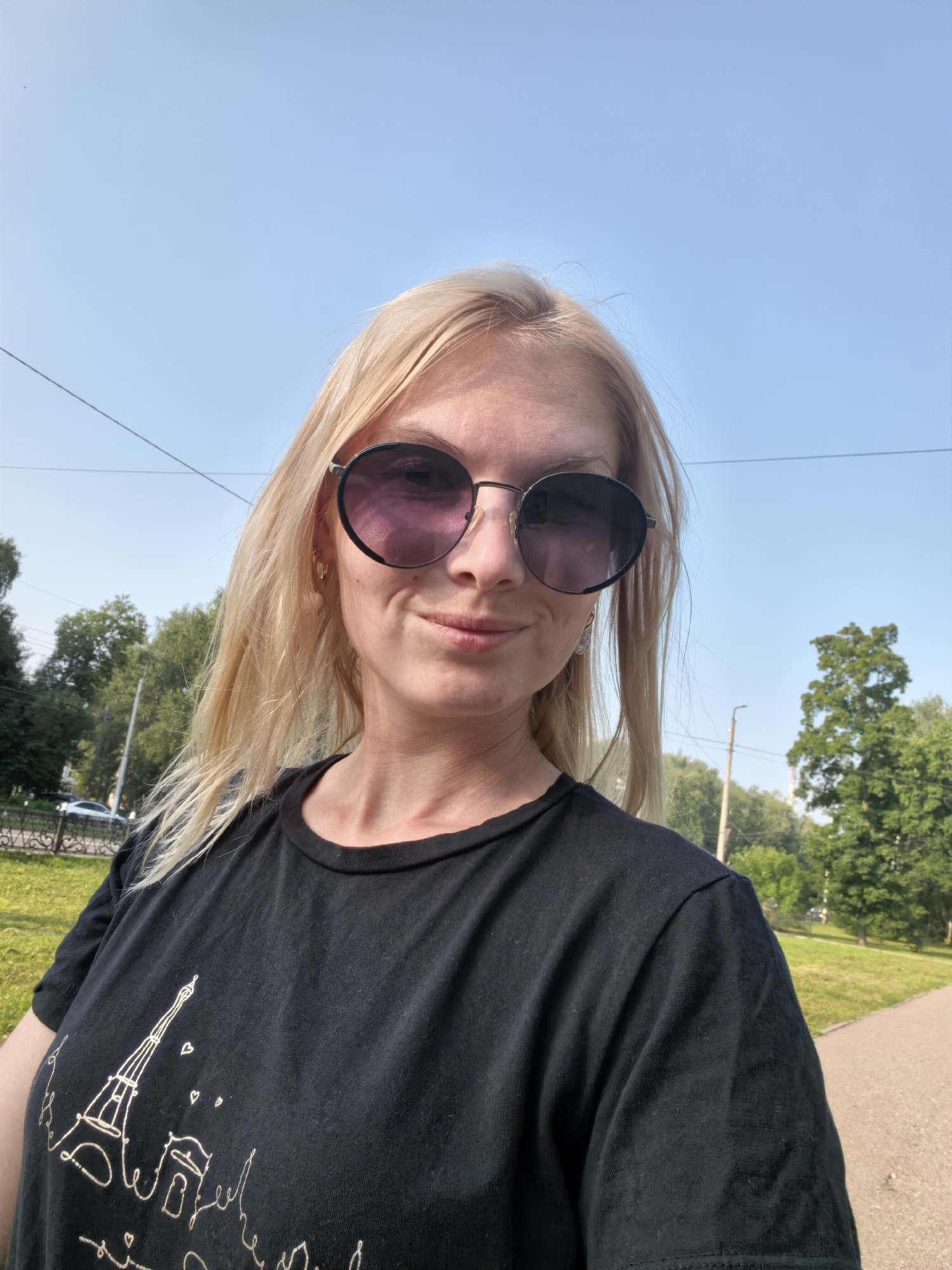 A blonde woman wearing sunglasses and a black t-shirt with a Paris-themed graphic is taking a selfie outdoors on a sunny day. The background includes green trees, a paved path, and a clear blue sky. This scene is likely in a park or suburban area of Sterlitamak, Russia, given the presence of greenery and what appears to be a public walkway.