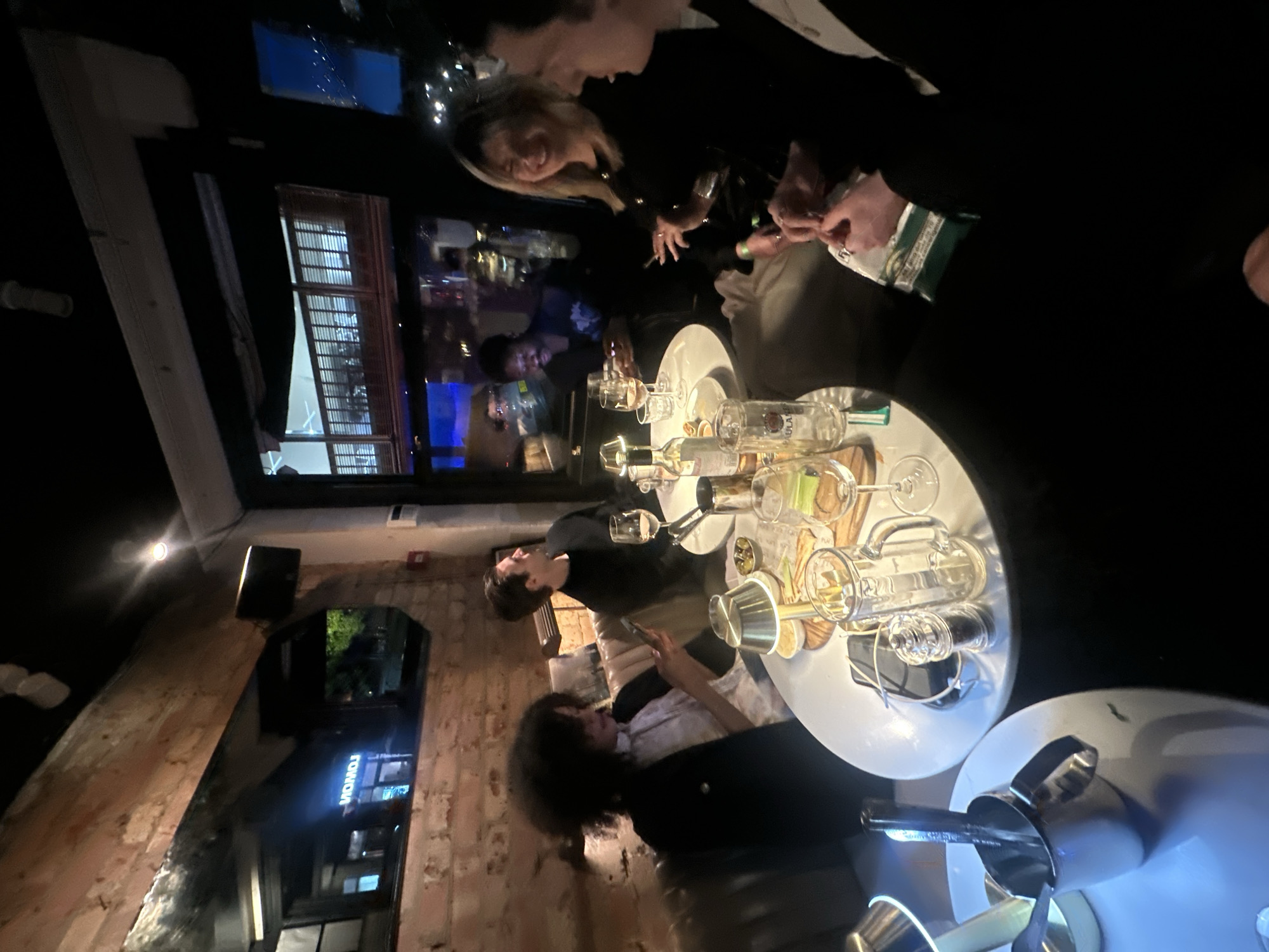 Here's a description of the image:
**Scene:** An indoor shot taken in what appears to be a bar or restaurant setting.
**People:** Multiple individuals are visible, including groups seated around tables. Some are dressed in dark clothing, possibly formal attire. Some are engaged in conversation.
**Objects:** Tables are prominent, laden with drinks, glassware, and possibly food items. Other objects include a bar or restaurant, windows overlooking the city.
**Environment:** The setting appears to be in London, United Kingdom. The interior features a modern design with brickwork. The overall atmosphere is of a lively, social gathering.
