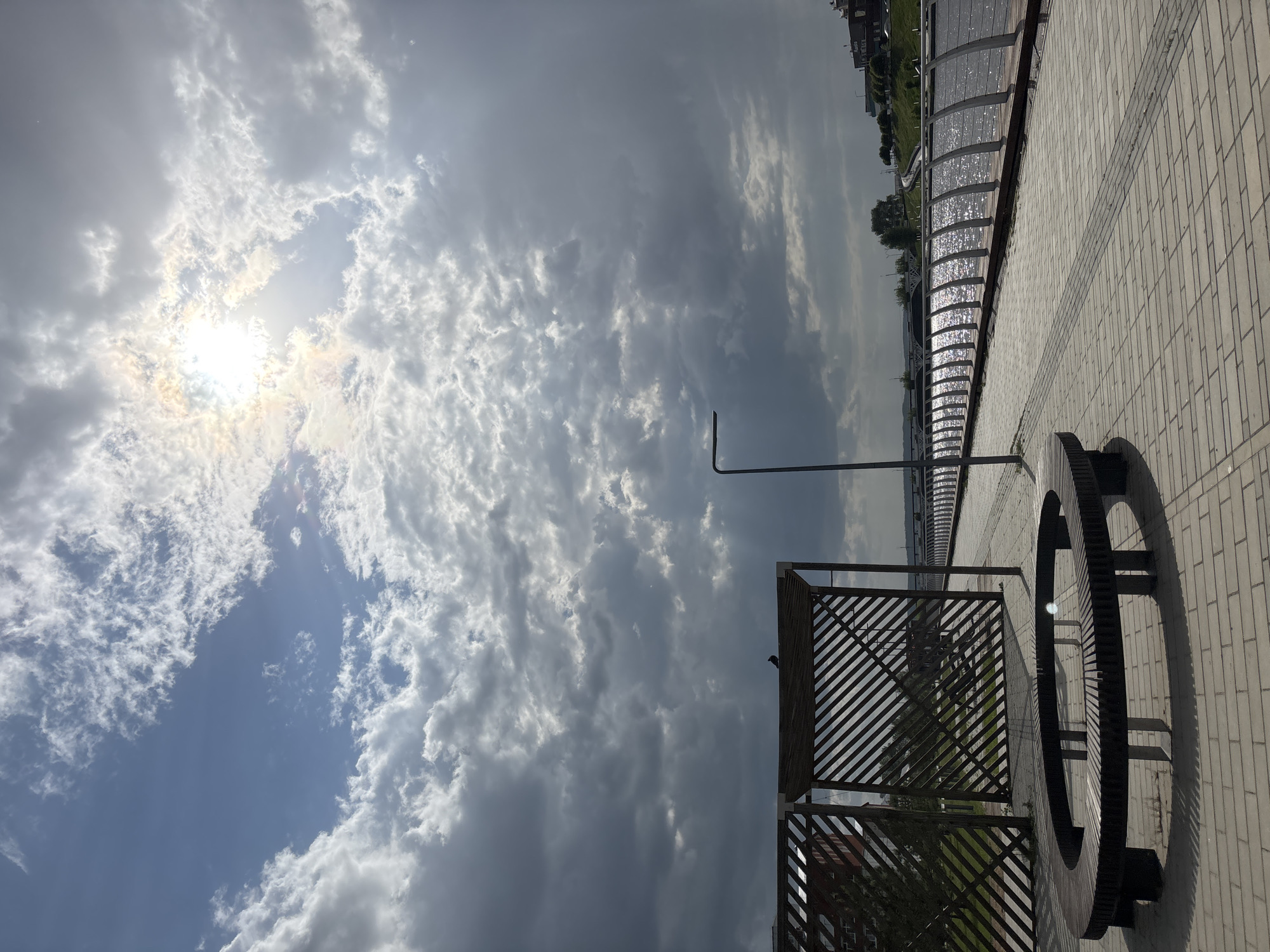 The image shows a paved walkway in Votkinsk, Russia, under a partly cloudy sky with the sun visible through the clouds. The walkway is made of light-colored rectangular bricks. To the right, there's a series of curved, glass-covered structures, possibly shelters or decorative elements, reflecting sunlight. A large, circular bench or installation is positioned on the walkway. A metal structure with a lattice pattern stands near the bench. A street light pole is visible against the sky. The perspective is tilted, making the sky appear to be above and to the left.