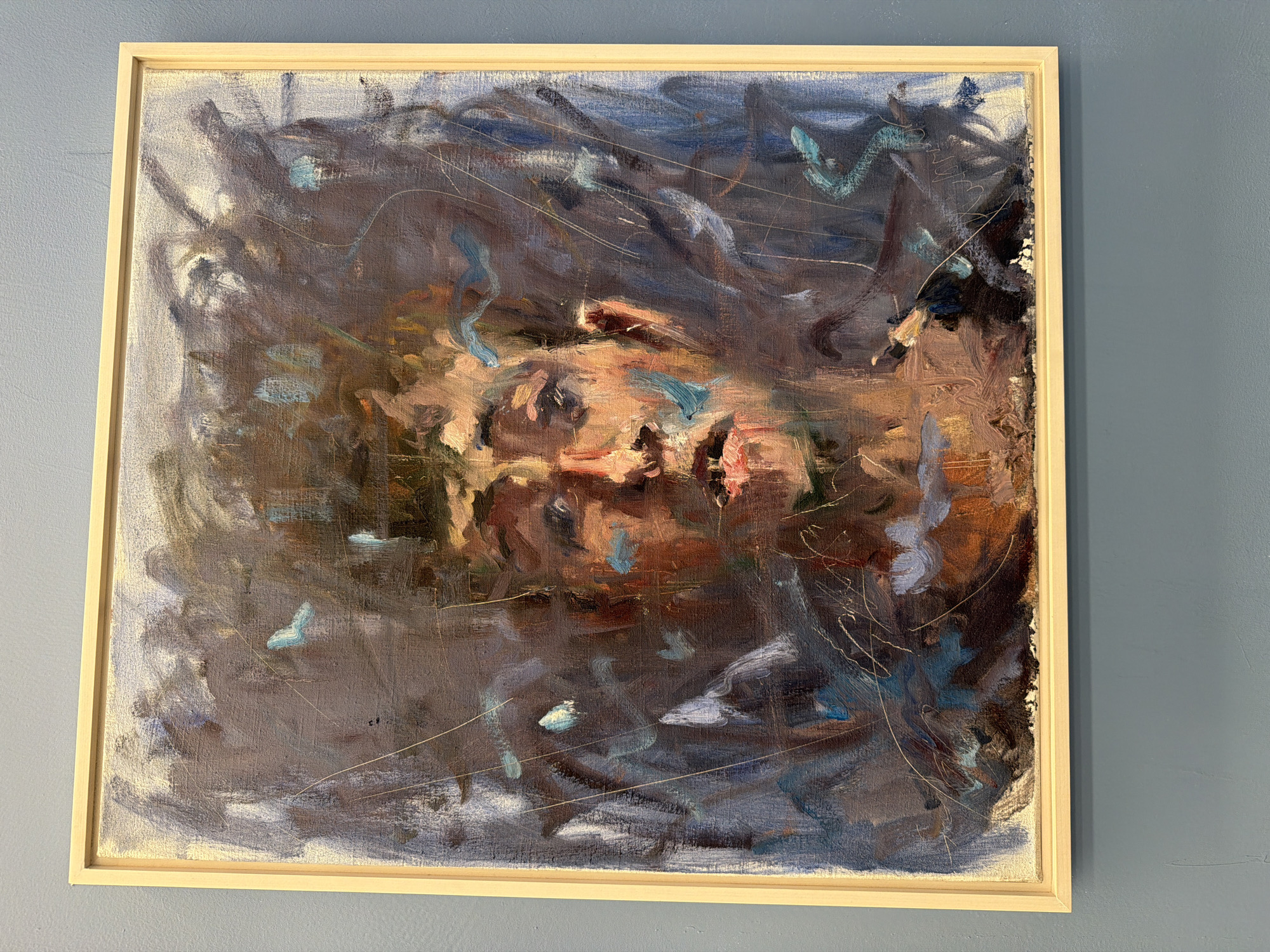This is an abstract oil painting framed in a light wood frame. The painting features a canvas of swirling, energetic brushstrokes in dark blues, purples, browns, and some touches of light blue and pink. The central area of the painting appears to hint at a human face or figure, with prominent use of peachy and brown tones. Delicate white and yellow lines are scribbled across the canvas, adding a sense of texture and movement. The overall impression is one of intense emotion, perhaps turmoil or introspection, rendered in a dynamic and expressive style. There are no discernible objects or specific setting details beyond the abstract representation. The background is a plain, muted blue-grey wall, consistent with an indoor gallery or exhibition space in Tel Aviv, Israel. No visible text is present on the artwork itself.