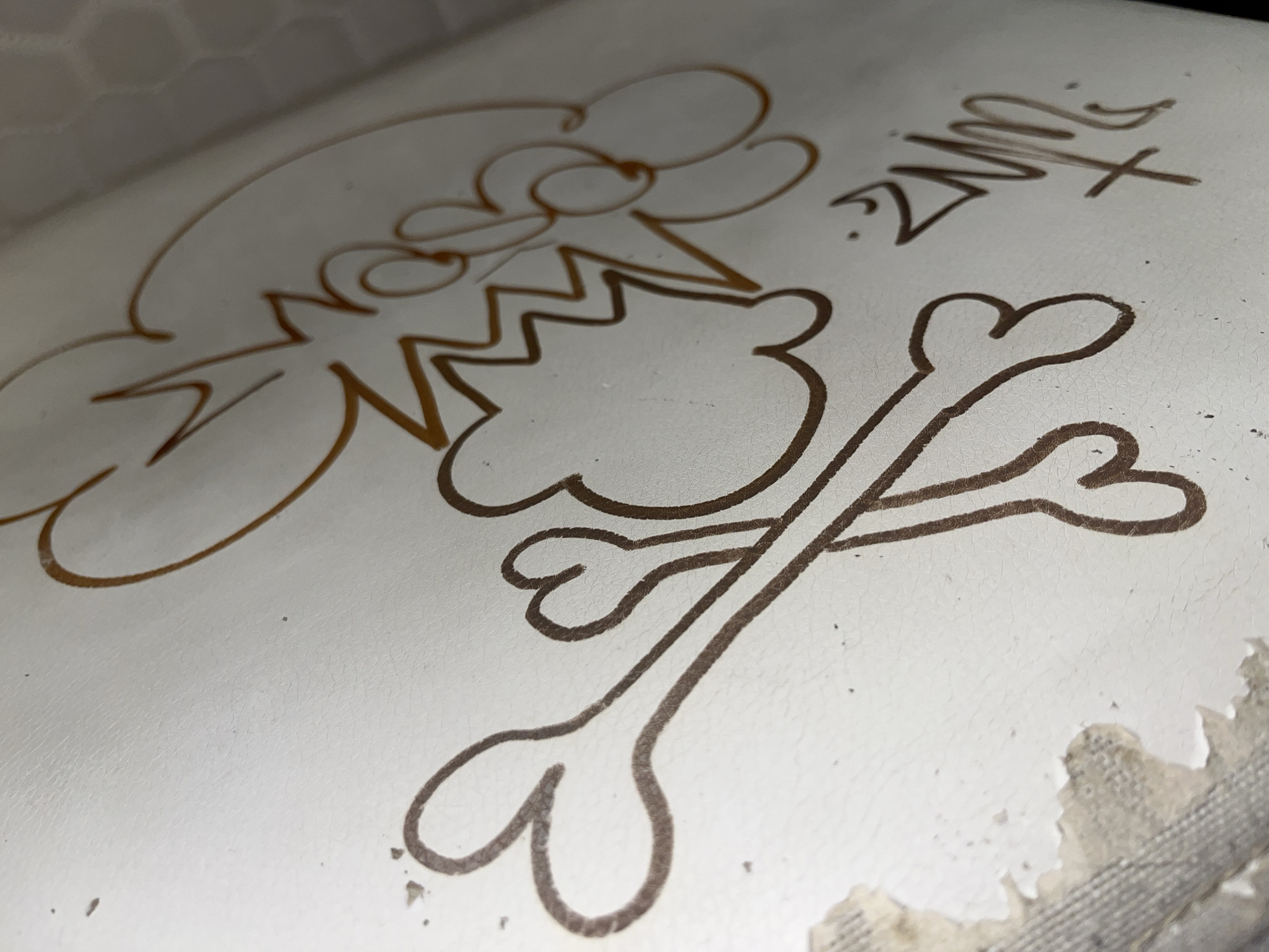 An overhead close-up shot shows a white surface with a brown drawing on it. The drawing is in the style of a cartoon, with bold brown outlines. On the left side of the image, there is a large, abstract shape resembling a cloud or a stylized face with jagged lines inside it. To the right of this shape, two crossed bones are drawn. The bones are in a simple, cartoonish style. In the upper right corner of the image, there is a signature that appears to read "Zune" in a stylized font, with a "+" symbol above the "e". The white surface appears to be somewhat textured and worn, with some areas of peeling material visible at the bottom right edge of the image. The overall impression is that of graffiti or a doodle on a white surface. The location of this object is Unknown, Unknown.
