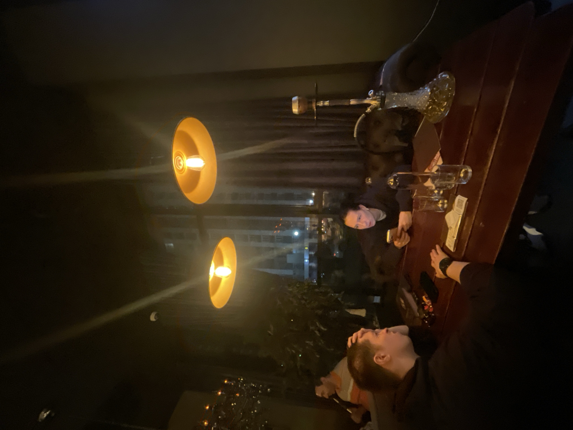 The image depicts a dimly lit interior scene, possibly a hookah lounge or restaurant, at night. In the foreground, two individuals are seated at a wooden table. The person on the left, a male with short hair and wearing a dark hoodie, has his hand on his forehead, appearing pensive or tired. On the table in front of him are some small objects, including what appear to be keys and a wallet. The person on the right, a female with dark hair, is looking at her phone. On the table between them are several glasses, a bottle of liquid, and a menu or papers. A hookah pipe with a decorative glass base is prominently displayed on the table to the right. In the background, another individual is visible, partially obscured, wearing an orange and white striped shirt and holding a hookah hose. Behind them, a window shows a blurred cityscape with illuminated buildings, suggesting an urban environment. The overall lighting is warm and ambient, provided by overhead pendant lamps. The atmosphere suggests a relaxed or social gathering. There is no information in the image to indicate a specific location within Krasnoyarsk, Russia.