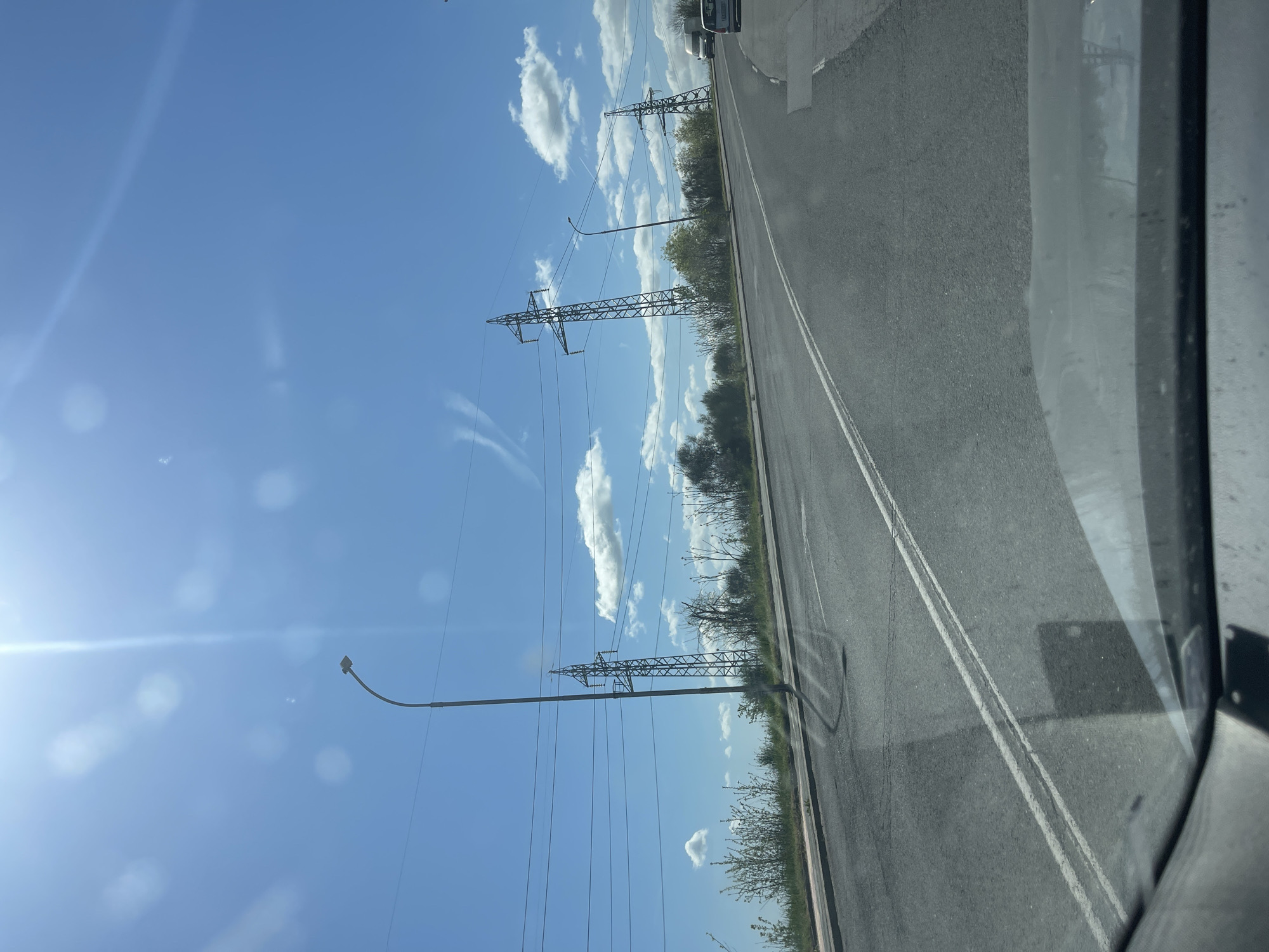 The image captures a bright, sunny daytime scene from the interior of a moving vehicle. A paved asphalt road, featuring a double white line marking the center, runs diagonally from the mid-right towards the upper right of the frame. Alongside the road, a strip of green vegetation, including bushes and small trees, extends into the distance.
Dominating the left side of the scene are several tall metal lattice transmission towers (electricity pylons), which support numerous power lines stretching horizontally across the clear blue sky. A single street light with a dark, curved arm and a rectangular fixture is visible in the lower left quadrant. The sky is largely clear, dotted with fluffy white cumulus clouds. A strong sun in the upper left creates a distinct lens flare effect. The front windshield and a portion of the vehicle's side mirror are visible on the far right, framing the view. Another vehicle can be seen in the distance on the road. The overall setting is an open area, likely on the outskirts of a city or along a highway, consistent with a location like Burgas, Bulgaria. There is no visible text in the image.