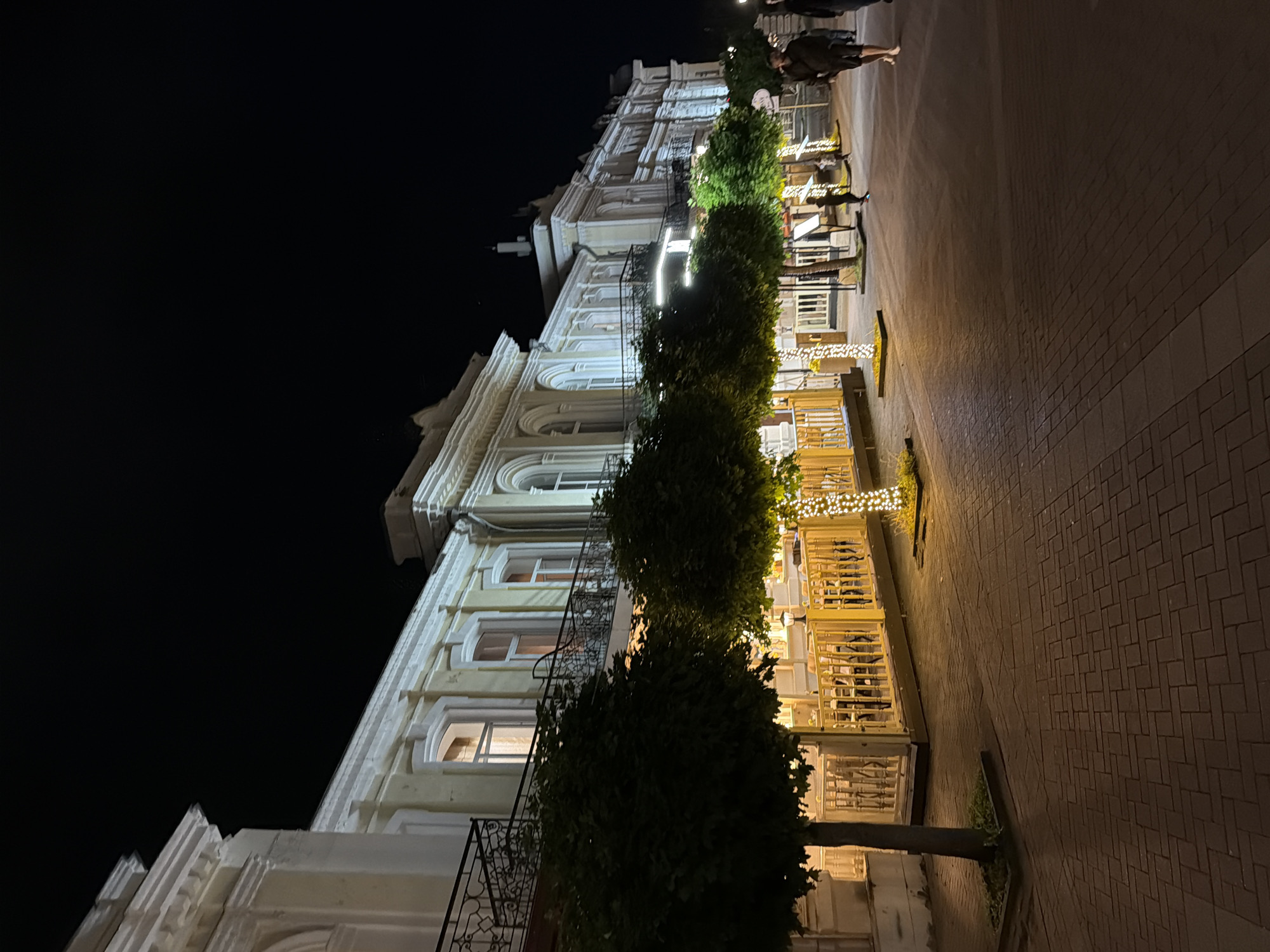 The image depicts a street scene at night in Kislovodsk, Russia, likely on a pedestrian walkway or promenade adjacent to a building that appears to be a hotel or a historic structure. The building, with its light-colored facade and ornate architectural details, is illuminated, highlighting its classical style with arched windows and decorative cornices. A balcony with intricate metal railings runs along the upper level of the building.
In the foreground, there is a row of trees, some of which are decorated with string lights, creating a festive atmosphere. These trees are pruned into a rounded shape. Adjacent to the trees is a raised outdoor seating area, possibly a cafe or restaurant terrace, with yellow railings and tables and chairs visible within. The ground is paved with brickwork, and the scene is lit by streetlights and the lights from the building and decorations.
Several people are visible in the scene. On the right side, two individuals are walking away from the camera down the paved pathway. Further back, near the outdoor seating area, a few more figures are present, including a child who appears to be running. The overall ambiance suggests a lively evening in a well-maintained urban environment. The dark sky indicates it is nighttime.