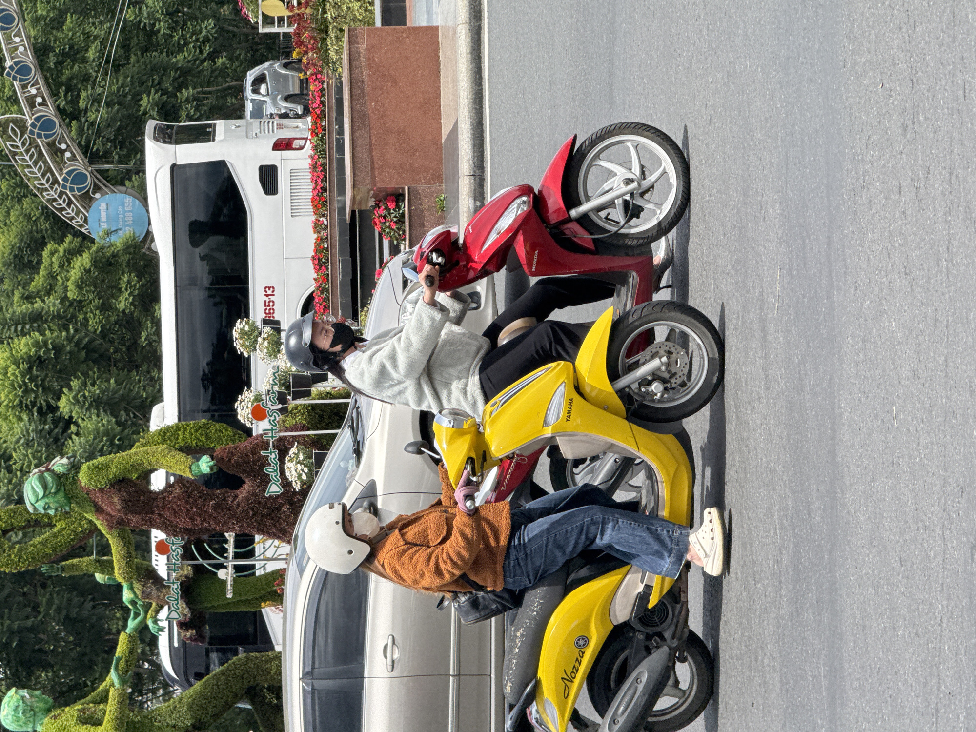 Two women are riding motorcycles in Da Lat, Vietnam. The woman in the foreground is on a yellow scooter, wearing a rust-colored jacket, blue jeans, and white Crocs. She has a white helmet and a light-colored face mask. The woman behind her is on a red scooter, wearing a grey sweatshirt, black pants, and black heels. She has a black helmet and a black face mask. In the background, there are two large green topiary figures, a white bus, and a silver SUV. The scene appears to be on a street with trees and flowers lining the sides. The bus has the number "B6513" on its side. The yellow scooter has "NOZZA" and "YAMAHA" written on it. The red scooter has "HONDA" written on it.