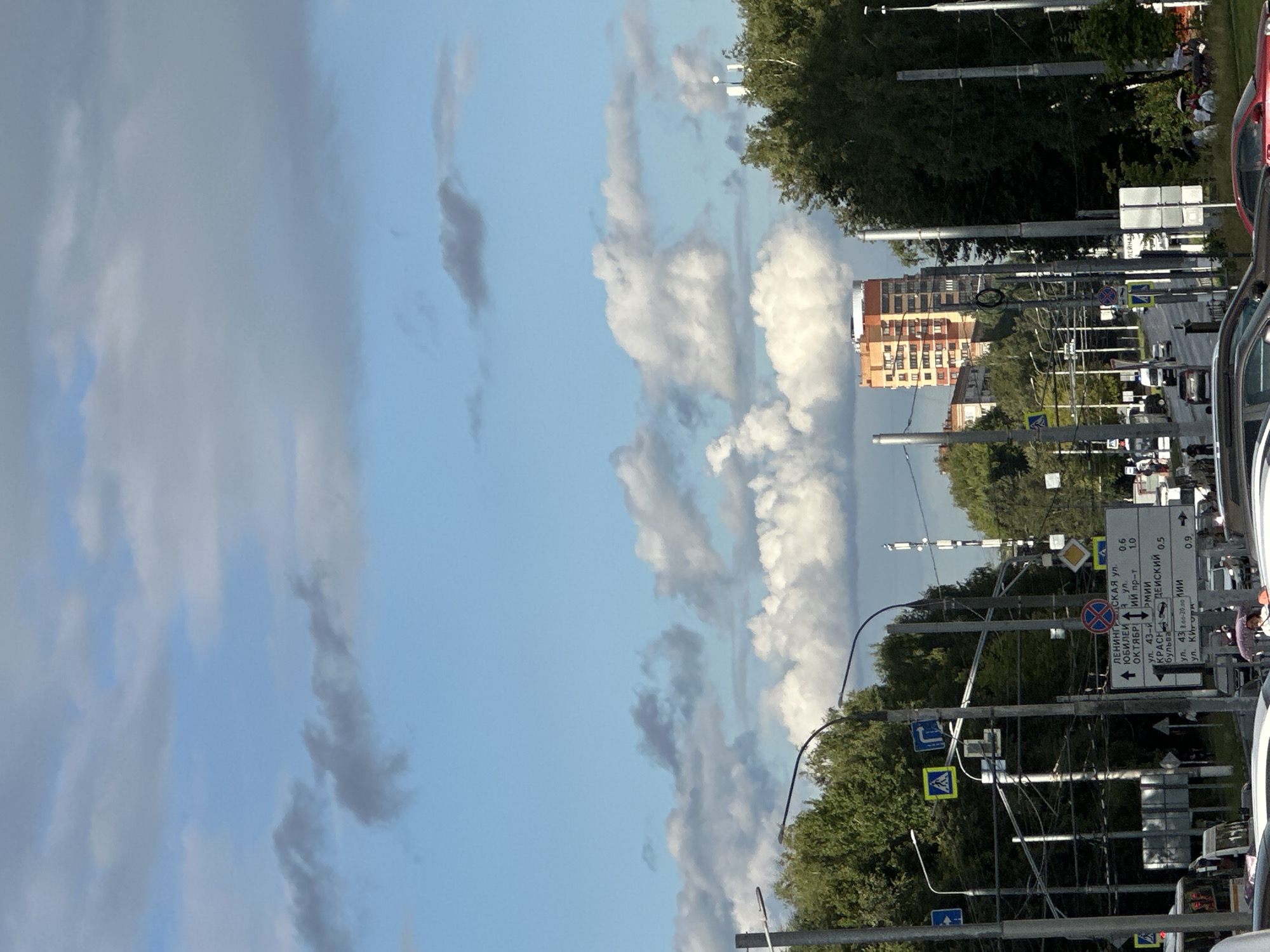 The image depicts a street scene in Podolsk, Russia, under a partly cloudy sky. In the foreground, several cars are visible on a road, with a white bus on the left. Streetlights and utility poles line the street, accompanied by various road signs, including pedestrian crossing signs and directional signs with Russian text. Tall green trees border the road on both sides, and a multi-story orange and beige building is prominent in the background. Several people are present, some walking and others gathered in small groups, with some holding umbrellas. A cell tower is also visible in the distance. The overall impression is of a typical urban environment on a day with mixed weather conditions.