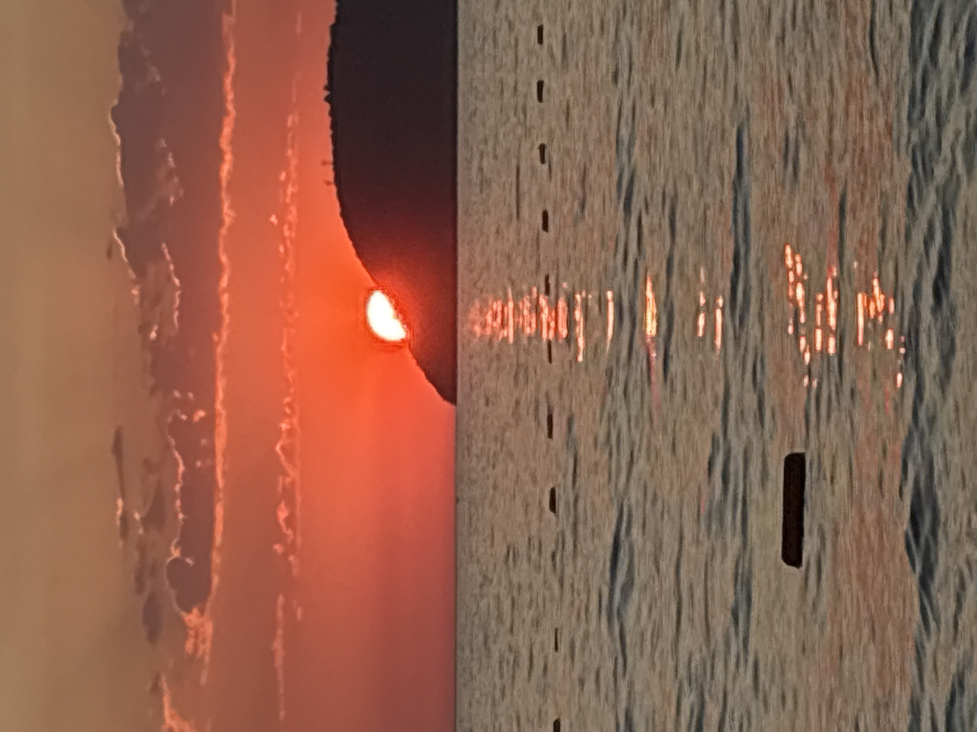 The image depicts a sunset over the Adriatic Sea, likely near the city of Bar, Montenegro. The sun, a vibrant orb of orange and red, is partially obscured by a dark, silhouetted hill or island on the horizon. The sky is filled with a gradient of warm colors, from deep orange near the sun to a lighter peach and pink higher up, with wispy clouds catching the fading light. The sea is calm, with gentle ripples reflecting the fiery hues of the sunset. A series of dark, rectangular buoys or markers are visible in a line across the water, suggesting a swimming area or demarcation. The overall impression is one of tranquility and natural beauty at the end of the day.