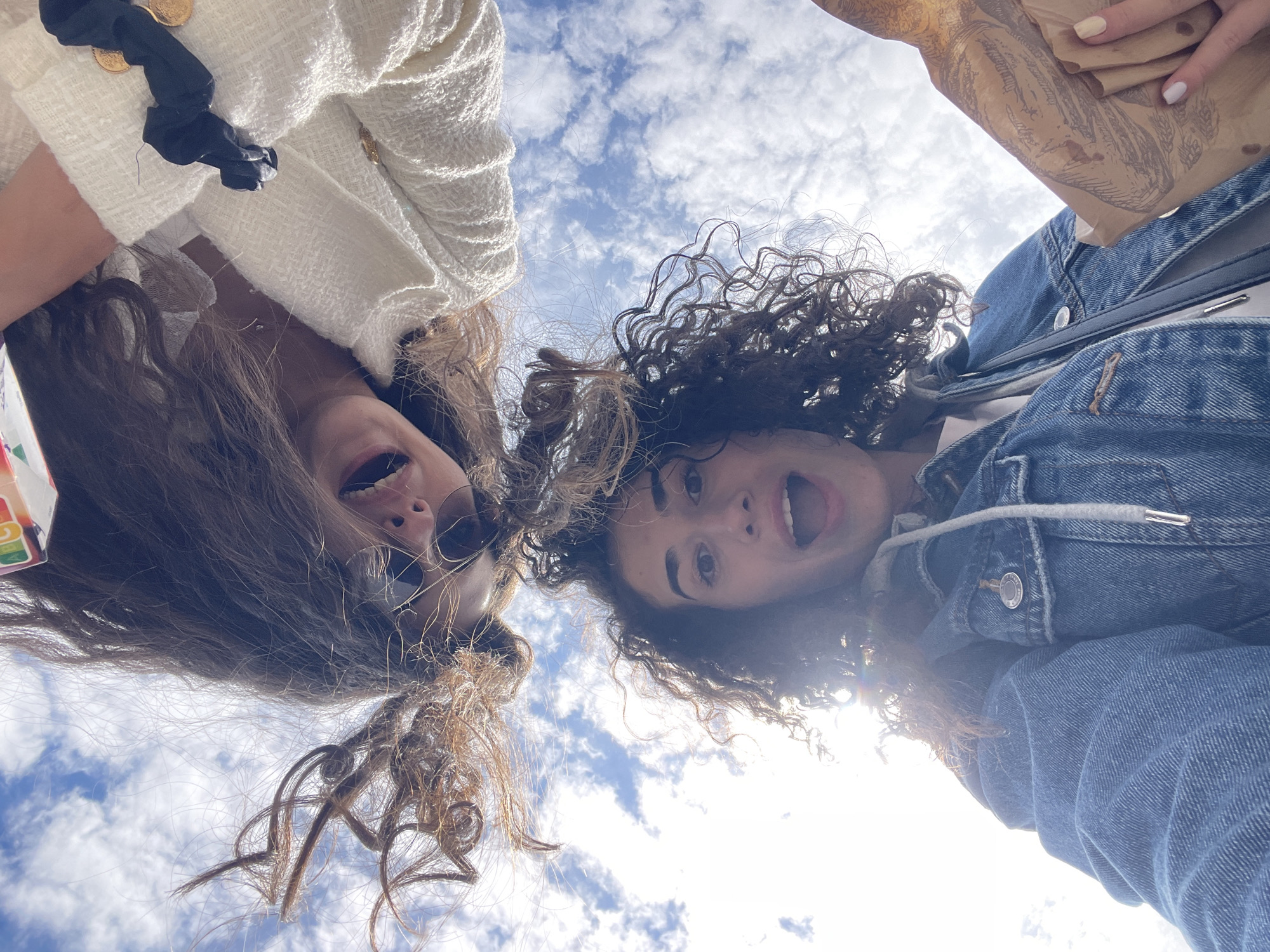 Two women are in the frame, positioned close together and shot from a low angle, looking down at the camera. The woman on the left has light brown curly hair and is wearing sunglasses, a white tweed jacket with gold button details and a black bow. The woman on the right has dark curly hair and is wearing a denim jacket. She's also holding a paper bag in her right hand. The background is a blue sky with scattered white clouds. The location appears to be outdoors. Information about Sofia, Bulgaria, is not discernible from the visual elements of this image.