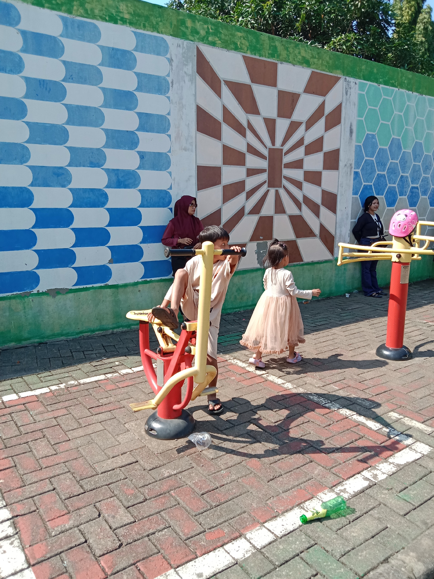 An outdoor scene in Kendal, Indonesia features a wall with colorful geometric murals, including blue and white ovals, a brown and white radial pattern, and light blue and green hexagons. In the foreground, children and adults are present. A young boy in a beige outfit is on a red and yellow exercise machine. A young girl in a light pink dress stands facing away from the camera. To the right, a woman in a black jacket and blue jeans stands near a red and yellow exercise machine topped with a pink helmet. Another woman in a maroon hijab stands by the blue and white section of the wall. The ground is paved with red and gray bricks. A green plastic bottle and a clear plastic bottle lie on the ground.