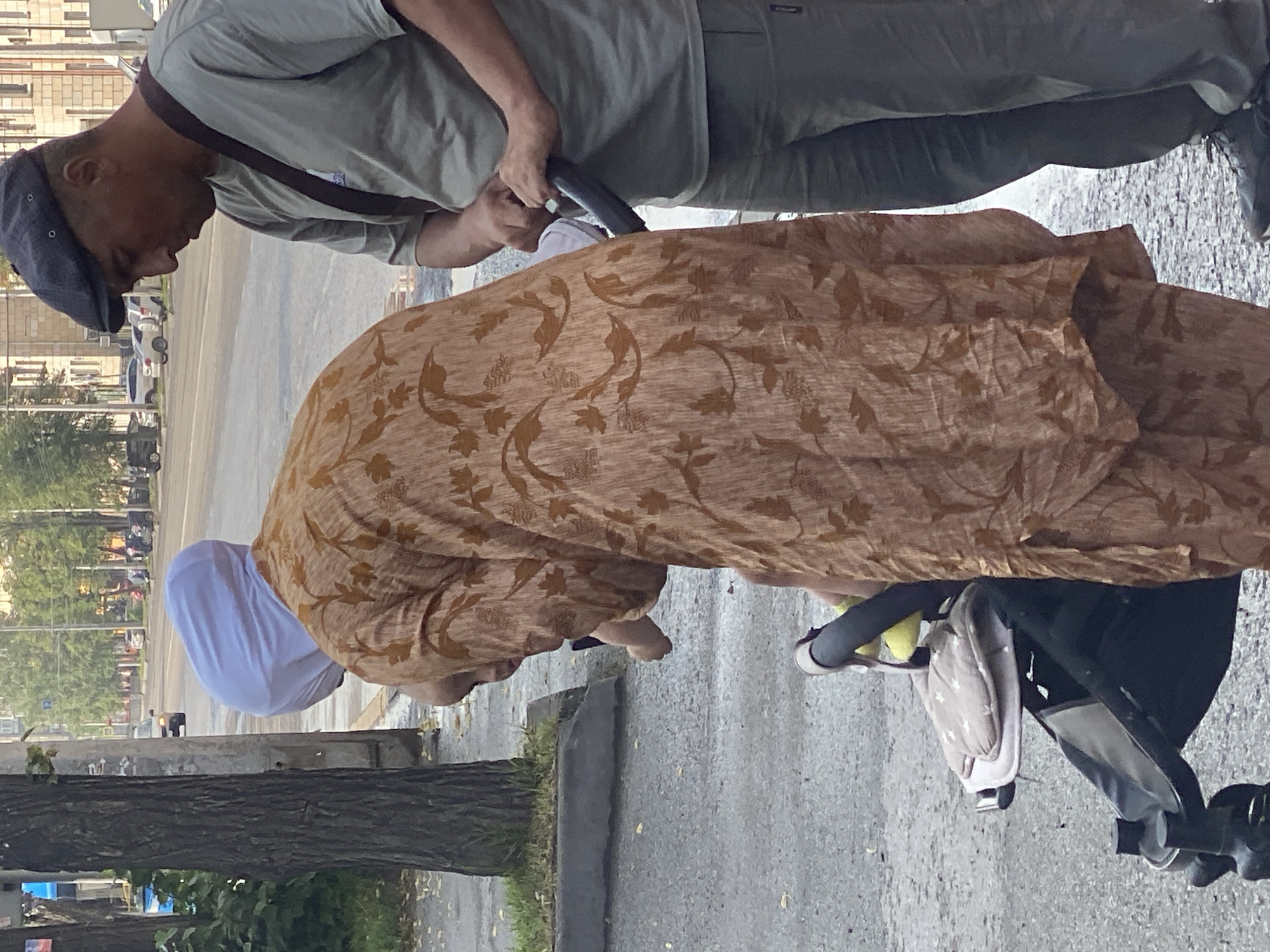 The image shows a street scene in Novokuznetsk, Russia. Two adults and a baby are near a baby stroller. A woman, partially visible, is wearing a tan, floral-patterned dress and a white headscarf. She is leaning over the stroller, which is black and has a white and gray patterned liner. A man, wearing a gray t-shirt, gray pants, and a dark flat cap, stands to the right of the woman. He is holding the stroller handle and looking down. The baby is also leaning out of the stroller. The background shows a street with cars, buildings, and trees. The ground is wet, suggesting it may have recently rained.
