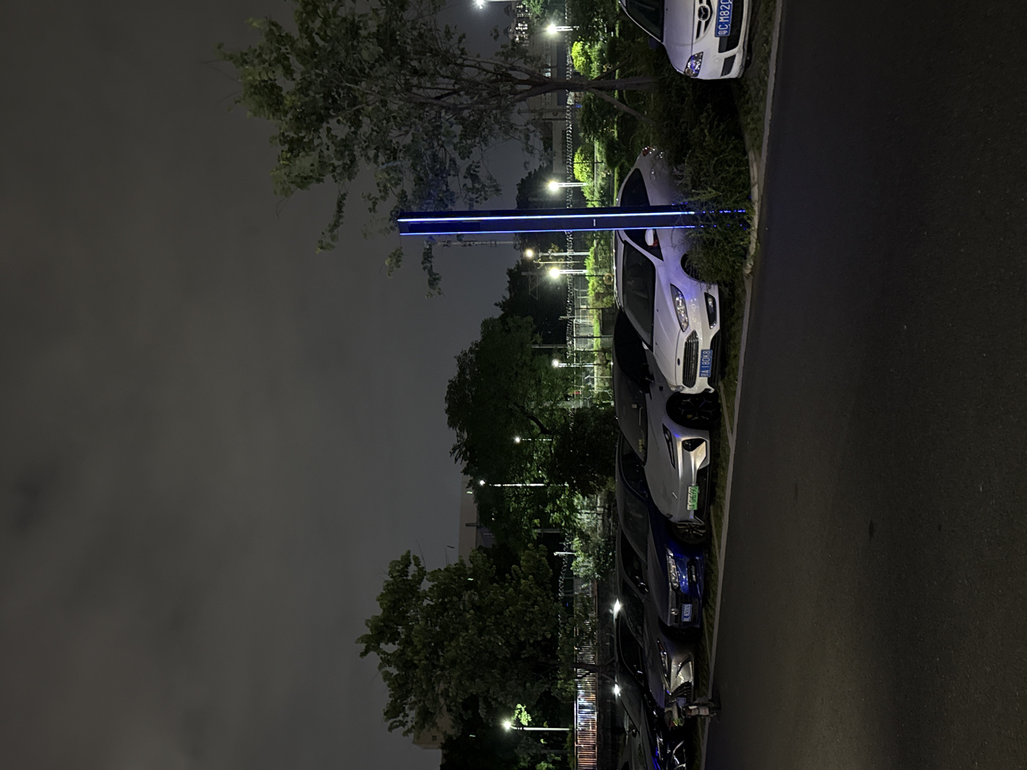 In Huangpu District, China, a row of parked cars is visible at night. The vehicles are arranged along a paved area, with a sidewalk and some vegetation bordering them. A tall, illuminated blue light pole stands to the right of the parked cars, casting a glow on the surrounding area. Trees and streetlights are scattered throughout the background, with a slightly overcast sky overhead. The scene suggests a public parking area in an urban or suburban setting.