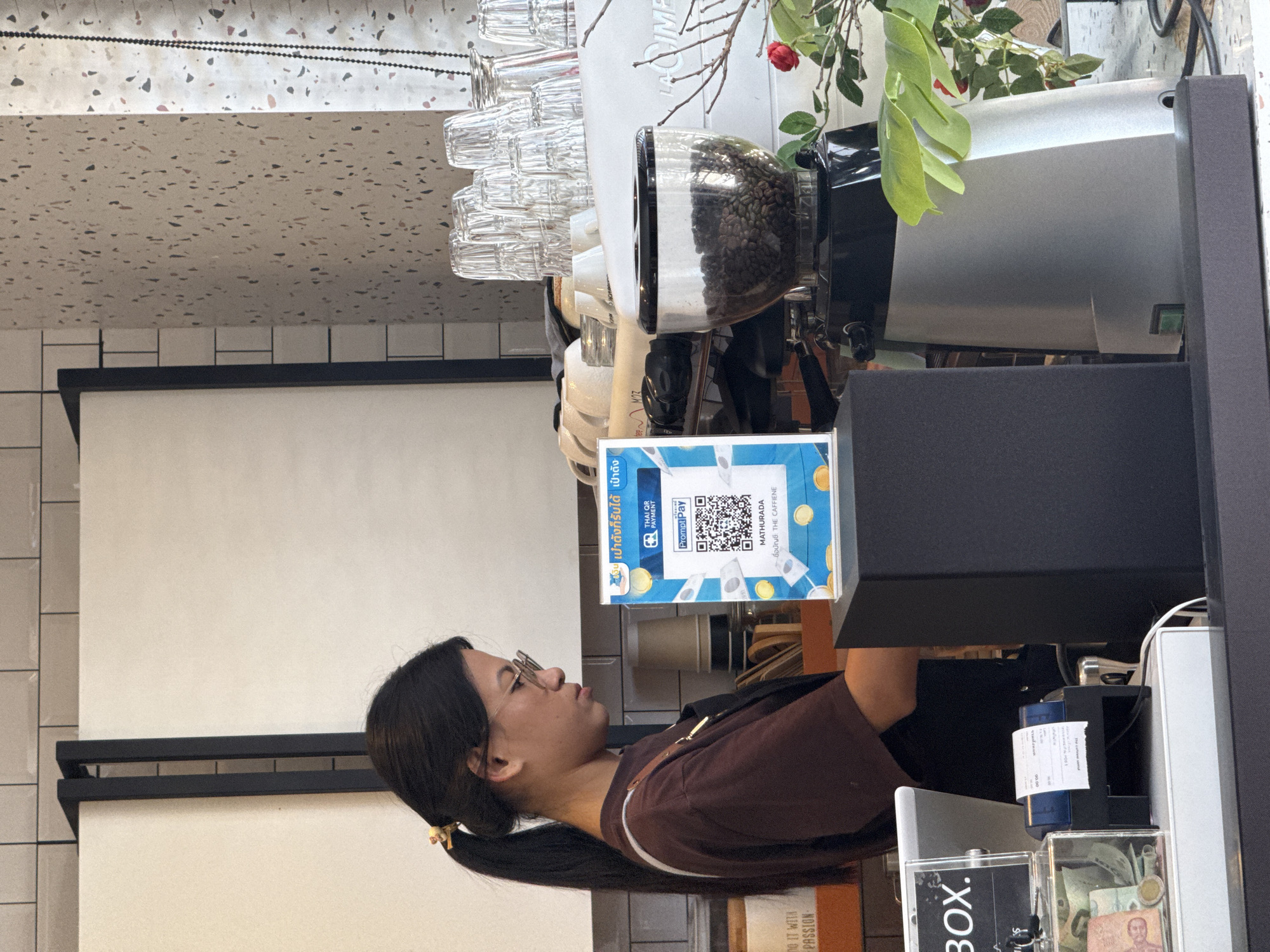 In Ko Samui District, Thailand, a young woman with dark hair pulled into a ponytail and wearing glasses is standing behind a counter in what appears to be a coffee shop. She is wearing a brown t-shirt and an apron. In front of her is a POS system displaying a QR code for PromptPay, a Thai payment system. To the left of the POS system is a clear plastic box containing Thai Baht currency. Behind her, there is a coffee grinder filled with coffee beans, stacked cups, and part of a white espresso machine. The background features tiled walls and a light-colored countertop. The overall scene depicts a typical retail transaction environment in Thailand.