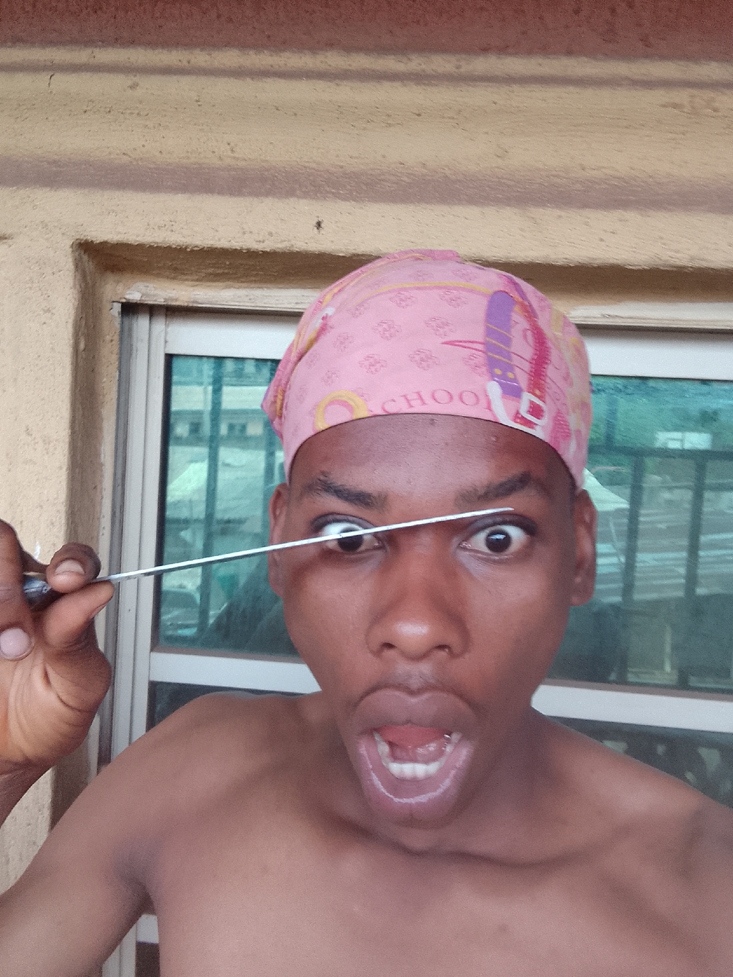 A bare-chested individual with dark skin is positioned in front of a window. The window frame is white, with the glass panes reflecting a blurred cityscape. The individual wears a pink head covering with a patterned design. A thin, metallic object resembling a skewer is held horizontally across the individual's left eye. The individual's mouth is open in an expression of surprise or shock. The background shows a building's exterior, painted in beige and tan. The location is Nkpor, Nigeria.