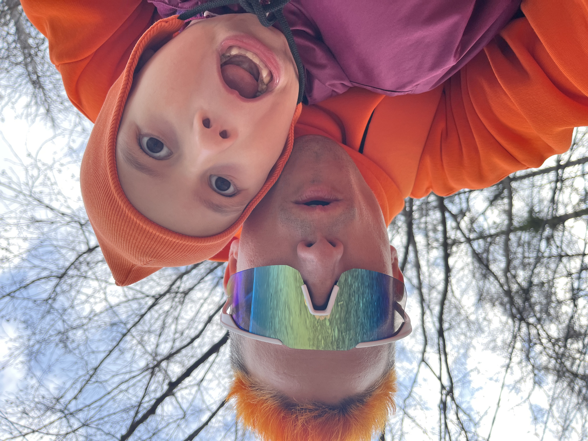 An upside-down, low-angle shot captures two individuals, an adult and a child, looking down towards the camera against a backdrop of bare tree branches and a bright sky. The perspective makes them appear as if they are hanging upside down.
The adult, positioned in the lower half of the frame (from the viewer's upright perspective), wears an orange hooded sweatshirt. Their hair, visible at the bottom of the frame, is dyed a vibrant orange. They wear large, futuristic white-framed sunglasses with iridescent, rainbow-colored mirrored lenses, reflecting the sky and branches. Their mouth is slightly open.
Above the adult, the child is also upside down, wearing an orange hooded sweatshirt and a purple top underneath. The child's mouth is wide open, showing teeth and tongue, and their eyes are looking directly at the camera, conveying a playful and energetic expression. A drawstring from the child's hoodie is visible, crossing between the two faces.
The background consists of a network of dark, bare tree branches against a bright, slightly overcast or hazy sky, suggesting a cold season like winter or early spring. The lighting is natural and bright, indicating daytime. The image captures a lighthearted moment, possibly a playful selfie or portrait taken outdoors in Chaikovsky, Russia. No visible text is present.