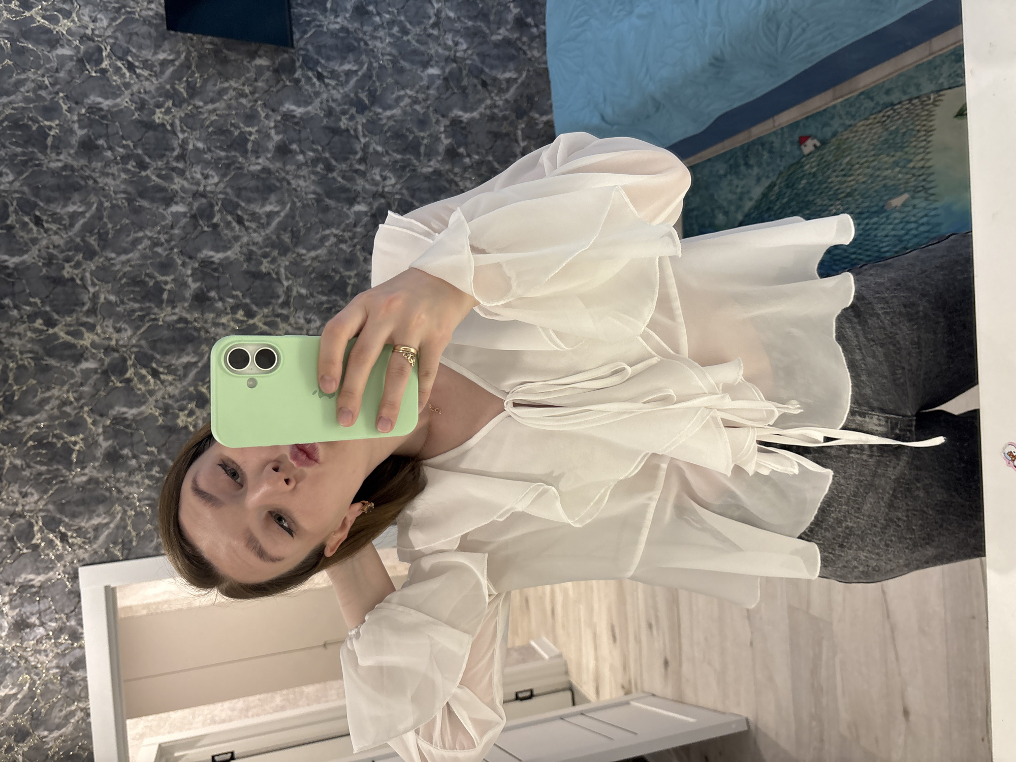 A young woman is captured in a mirror selfie, holding a light green iPhone with dual cameras in her right hand to take the picture. She is facing the camera with a slight pout, showcasing her makeup, including eyeliner and lipstick. Her brown hair frames her face.
She is dressed in a flowy, semi-sheer white blouse featuring prominent ruffles on the sleeves and along the front, tied with white strings. She wears dark gray bottoms, likely jeans or pants. Her right hand displays two gold rings, and a gold earring is partially visible on her right ear.
The background, seen as a reflection, features a dark, marble-patterned wall to the left and a lighter, possibly wooden or laminate floor. A blue and white patterned rug or tapestry with an abstract landscape, including a small red-roofed house, is visible in the upper right. White furniture or cabinetry and a white door frame are also reflected in the mirror.
The image portrays an indoor setting, likely a home or dressing room, in Izhevsk, Russia, with the woman posing for a self-portrait. There is no visible text in the image.