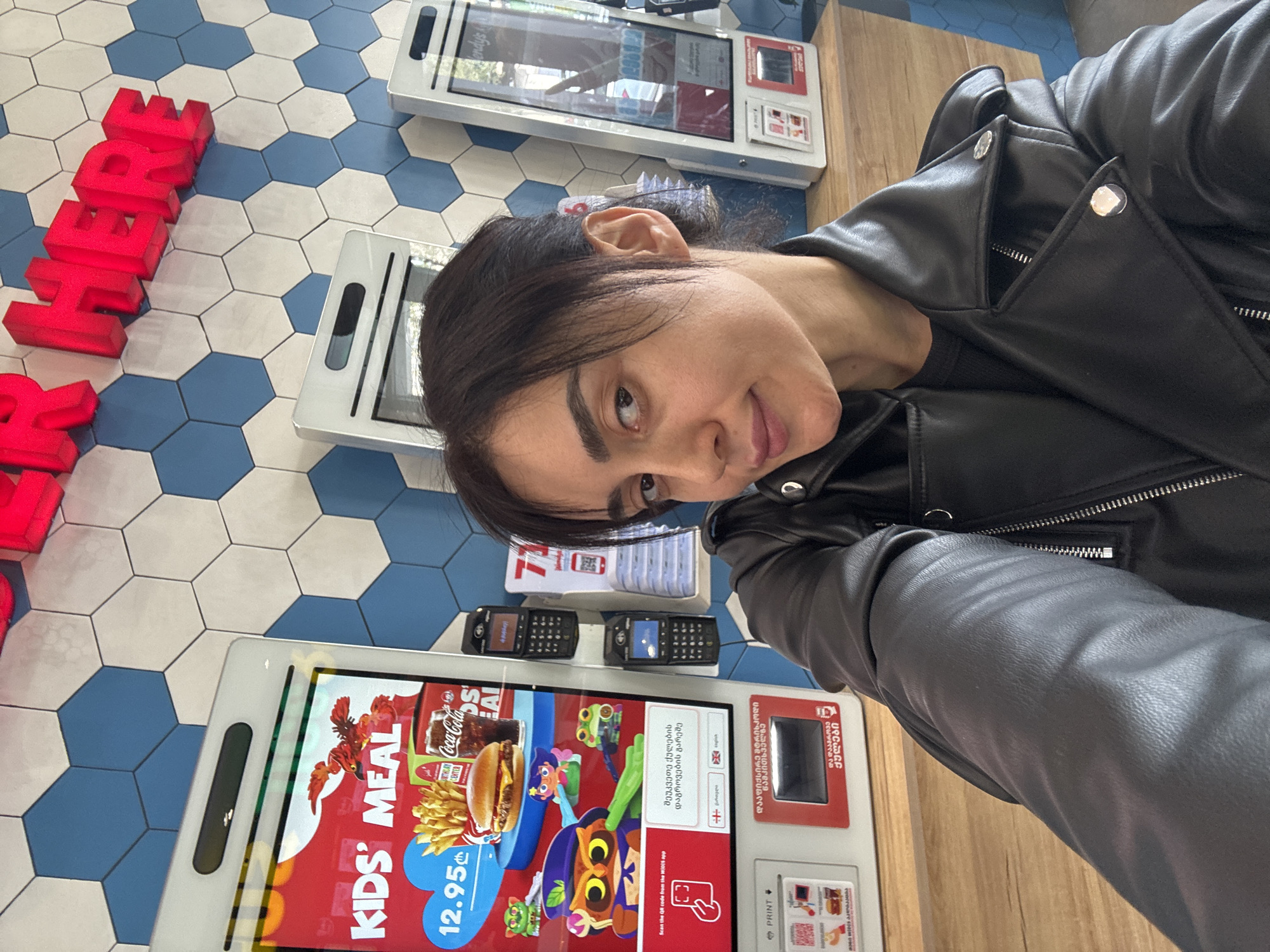 A woman wearing a black leather jacket is captured in a selfie taken in a fast-food restaurant, likely in Tbilisi, Georgia. She looks directly at the camera with a slight smile. Her jacket features silver buttons and zippers, and her dark hair is pulled back.
The setting is characterized by a modern self-service ordering area. To her left, large, red, illuminated letters spell out "HERE" on a hexagonal tiled floor of blue and white. Several self-service kiosks are visible, displaying menus and ordering options. The nearest kiosk showcases a "KIDS' MEAL" with a hamburger, fries, and Coca-Cola, priced at "12.95 ₾" (Georgian Lari). The screen also presents language choices including English, alongside text in Georgian script such as "შეუკვეთე ქულების დაგროვების გარეშე" (Order without accumulating points) and a "PRINT" button. A prompt to "Scan the QR code from the WIDIS app" is also visible. Between two of the kiosks, two small, black, older-model mobile phones are resting on a wooden surface. The presence of Georgian language and currency explicitly indicates the location.