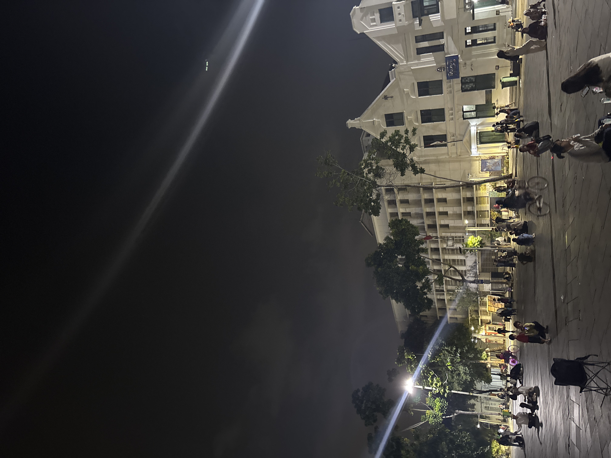 The image depicts an outdoor scene at night, likely in a city square or street. The dominant feature on the right side of the frame is a multi-story building with a white facade, characterized by rows of dark windows and some decorative architectural elements. A bright, diagonal light beam cuts across the dark sky on the left, originating from a light source outside the frame.
In front of the building, a group of people are gathered on a paved area. Many are seated or crouching on the ground, some appearing to be engaged in conversations or activities, while others are simply present. There are bicycles visible, suggesting a mode of transportation or perhaps part of the scene's atmosphere. The presence of trees with lush foliage interspersed between the building and the gathering area adds a natural element to the urban setting. Streetlights are also present, casting illumination on the area.
The time of day is clearly night, with the sky being dark and illuminated by artificial lights. The lighting suggests a clear or slightly hazy night. The overall mood of the scene is relaxed and communal, with people appearing to be enjoying a casual evening outdoors. The location is identified as West Jakarta, Indonesia. There is some visible text on a blue sign on the building, but it is not clearly legible.