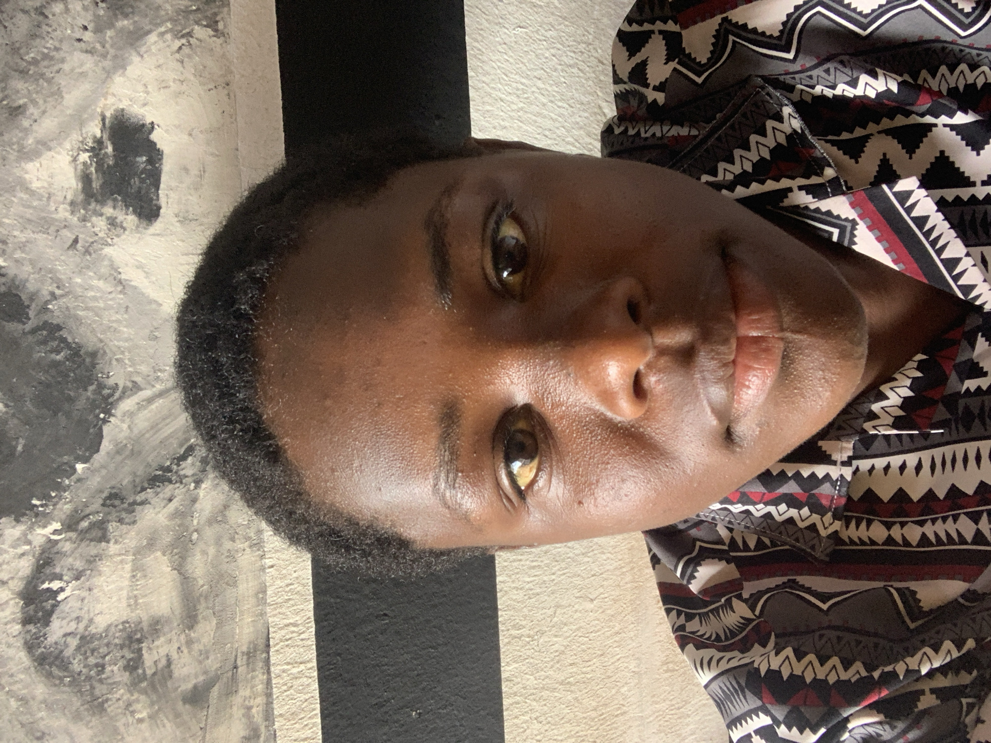 A close-up, tilted portrait shows a person with dark skin, brown eyes, and short black hair. The person is wearing a patterned shirt with black, white, grey, and red geometric designs. The background consists of a wall with both neutral and black painted sections. The location and city of Jos, Nigeria are not discernible from the image.