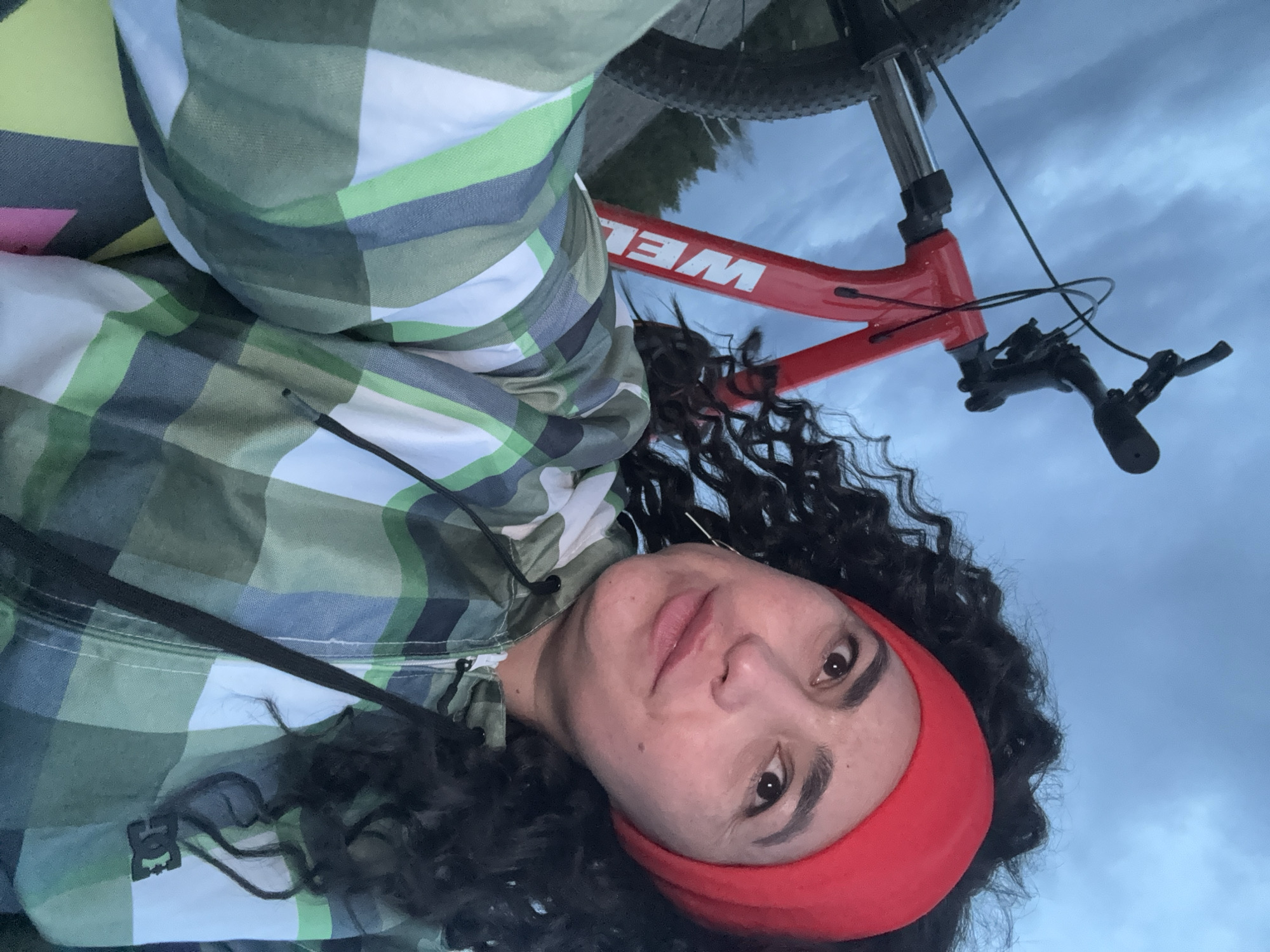 A selfie photograph taken in daylight shows a person with curly dark hair wearing a red headband and a plaid jacket with a DC logo. The person is positioned slightly to the left of the frame, looking at the camera. Behind the person is a red mountain bike with the word "WEL" visible on the frame, and the sky. The location is Кемуль, Russia.