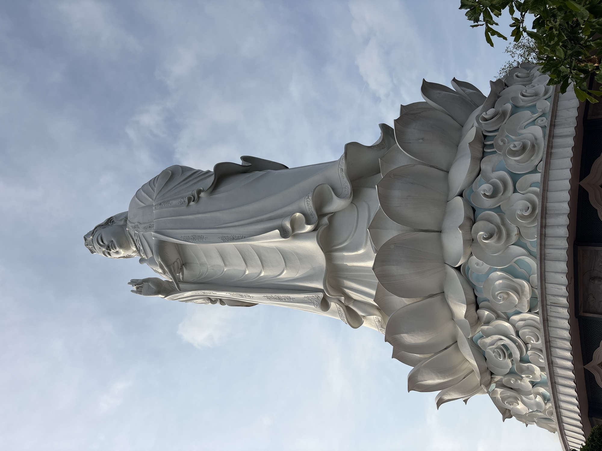 The image depicts a large, white statue of a Guanyin (Avalokiteśvara) Bodhisattva, viewed from a low angle against a cloudy blue sky. The statue is standing on a lotus flower pedestal with cloud designs, and some foliage is visible in the top right corner. This is the Lady Buddha statue at the Linh Ung Pagoda in Đà Nẵng, Vietnam.
