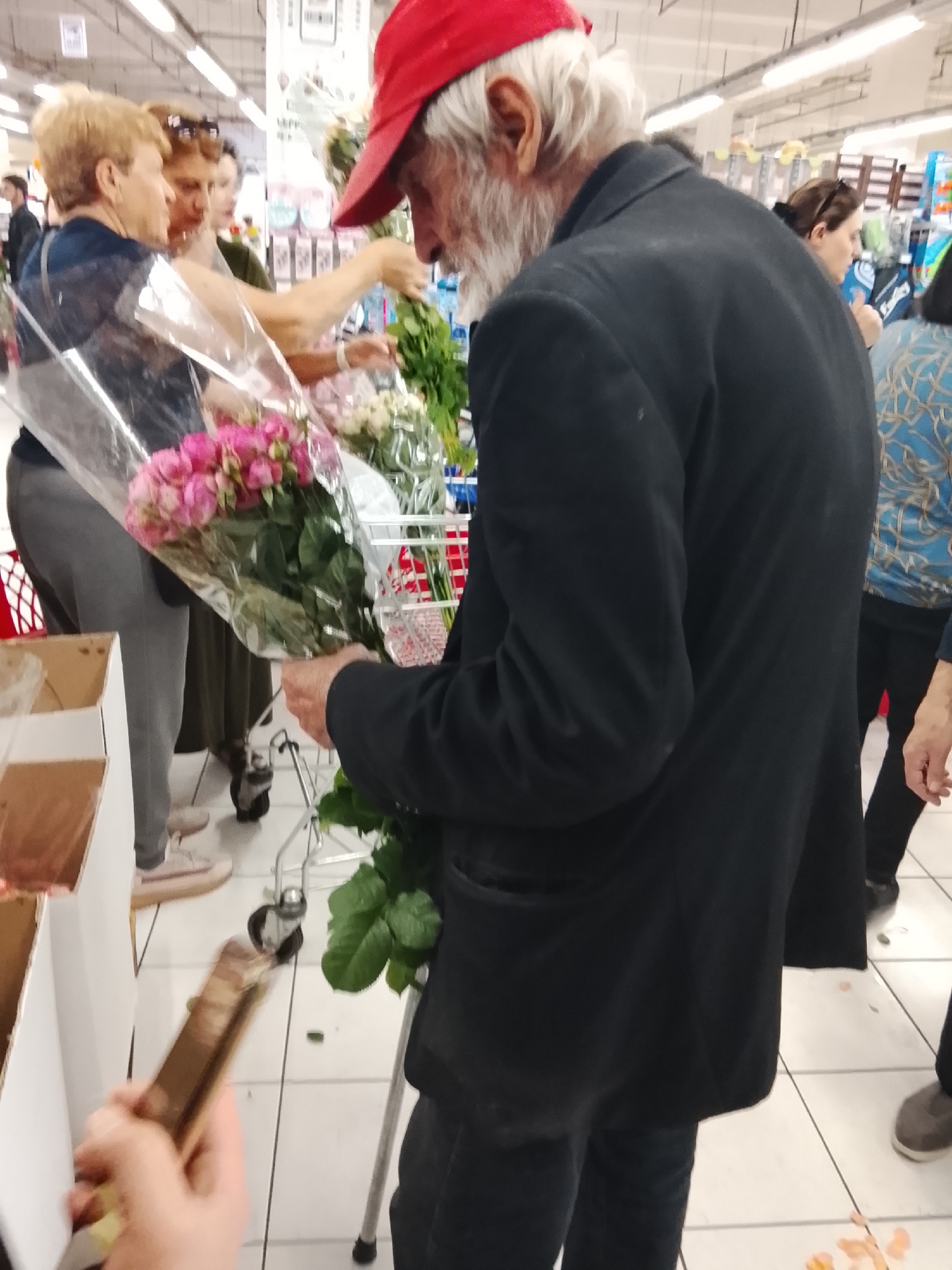 In an indoor environment that appears to be a supermarket, a Caucasian male with white hair and beard, wearing a red baseball cap and a black suit jacket, is seen from the rear, facing away from the camera. He is holding a bouquet of pink roses wrapped in plastic. In front of him, two Caucasian women are also present. One woman, with blonde hair, is wearing a blue shirt and grey pants. The other woman, with brown hair, is wearing sunglasses on her head and a dark-colored dress. The women are looking at the bouquet. The floor is tiled in a white grid pattern. Various floral displays and shopping carts are visible in the background, along with overhead fluorescent lighting. The scene suggests a shopping activity involving the selection of flowers.