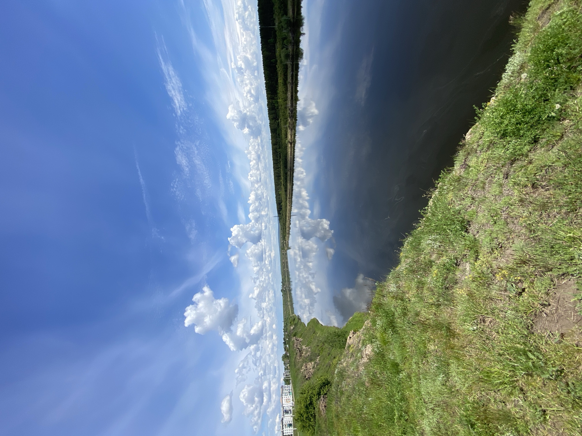 This image captures a vibrant, sunlit landscape, likely overlooking a wide river or lake in Novomaltinsk, Russia. The scene is divided vertically, with a bright blue sky on the left and the calm, dark water surface on the right.
The sky is adorned with various white clouds, including wispy cirrus and fluffy cumulus formations. These clouds are strikingly reflected in the water, creating a near-symmetrical mirrored image of the sky. A distant horizon runs vertically through the middle of the frame, where a dense line of dark green forest marks the opposite bank of the waterway. This forest is also reflected in the water's surface.
In the foreground, along the bottom edge of the image, a lush, green grassy bank slopes gently downwards towards the water. This bank is covered in verdant foliage and patches of exposed earth. In the far distance on the lower left, several white buildings are visible, suggesting a distant settlement or urban area.
The time of day appears to be midday or early afternoon, indicated by the bright sunlight and clear sky. The weather is sunny with scattered clouds, and there are no discernible activities or people present. No visible text is identifiable in the image. The overall impression is one of serene natural beauty and expansive open space.