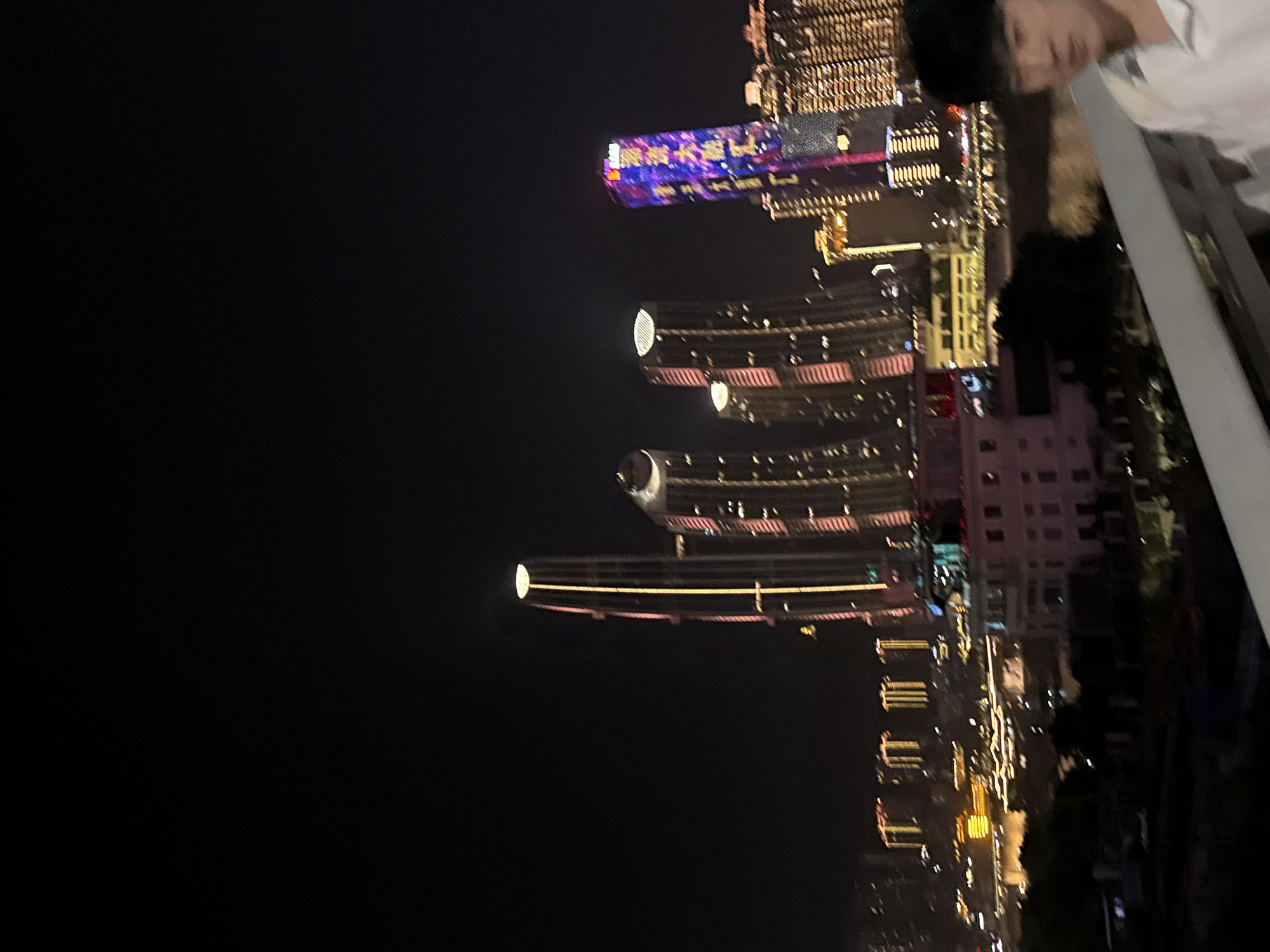 In Yuzhong District, China, at night, a wide shot captures a skyline dominated by illuminated skyscrapers. The perspective is from a slightly elevated position, possibly a bridge or balcony, looking towards the city. In the foreground, a portion of a white railing is visible. To the right of the frame, a young Asian male is partially visible, looking towards the camera. He is wearing a white collared shirt.
The background is a dense urban landscape featuring numerous modern buildings. Several tall skyscrapers stand out, their facades adorned with colorful lights. One prominent skyscraper on the left is tall and slender, with vertical lines of light. To its right, other skyscrapers are visible, some displaying pink and yellow lighting. Further to the right, a particularly striking skyscraper is covered in vibrant blue and purple lights, forming a dynamic pattern. Beyond these, more buildings with grid-like lighting extend into the distance. The overall ambiance is that of a modern, brightly lit city at night.