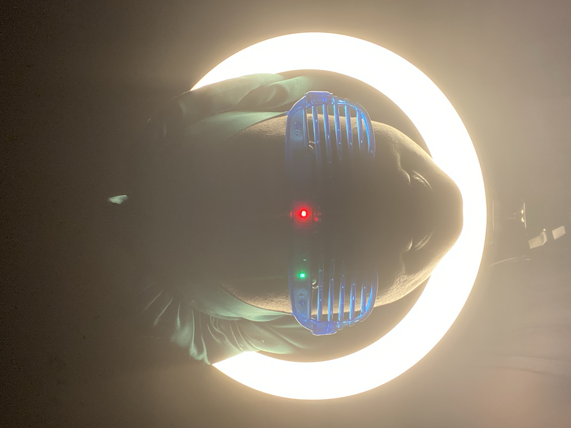 A person is lying on their back in what appears to be a dark environment. A ring light is positioned above their head, illuminating their face. The person is wearing blue plastic eyeglasses with a vertical, grid-like pattern. A red LED and a green LED are visible on the glasses. The person is wearing a green garment. The context of the location in Maigizo, Nigeria, is unknown.