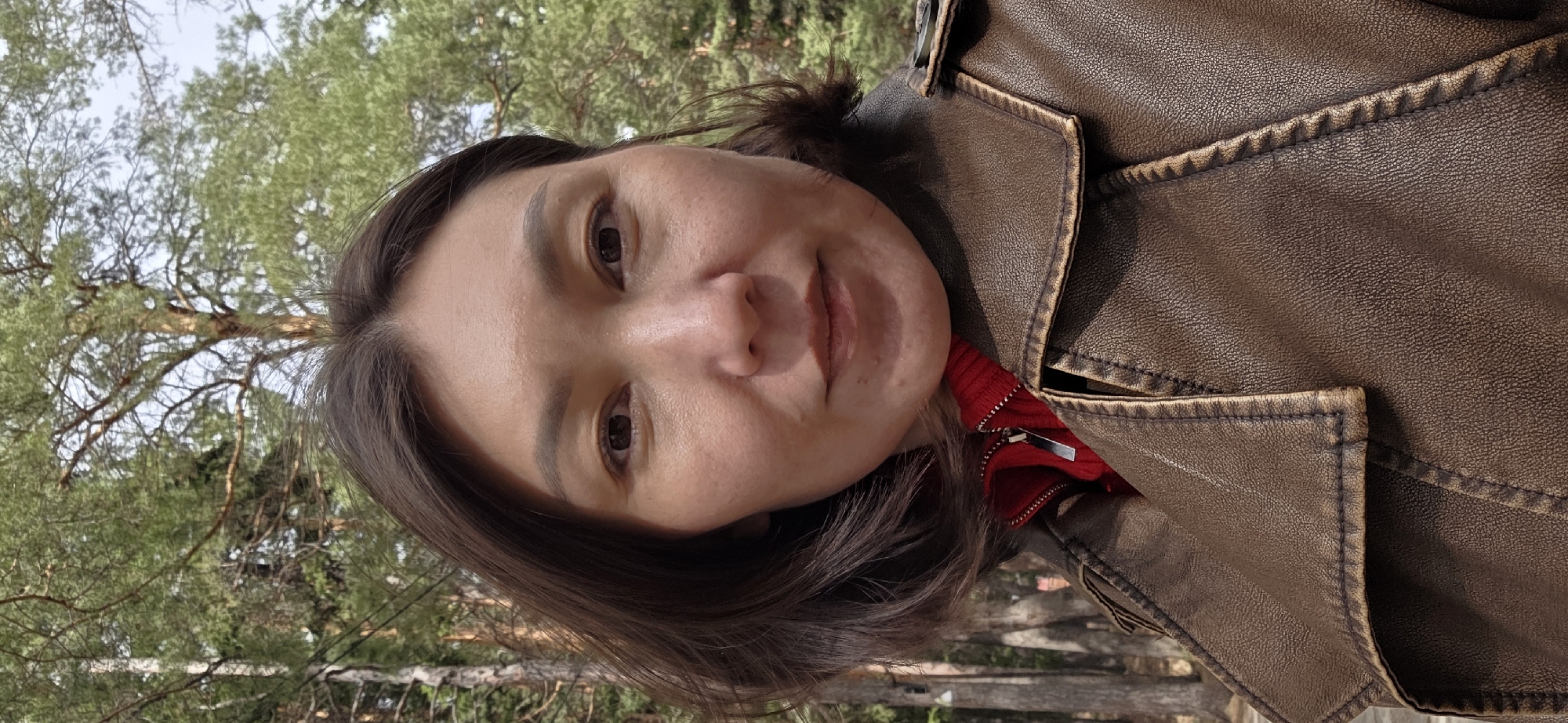 A woman with medium-length brown hair and fair skin is wearing a brown leather jacket with a red shirt underneath. The background appears to be a wooded area. While I can describe the image, I cannot provide a specific location such as Lensk, Russia, because there is no location information within the image.