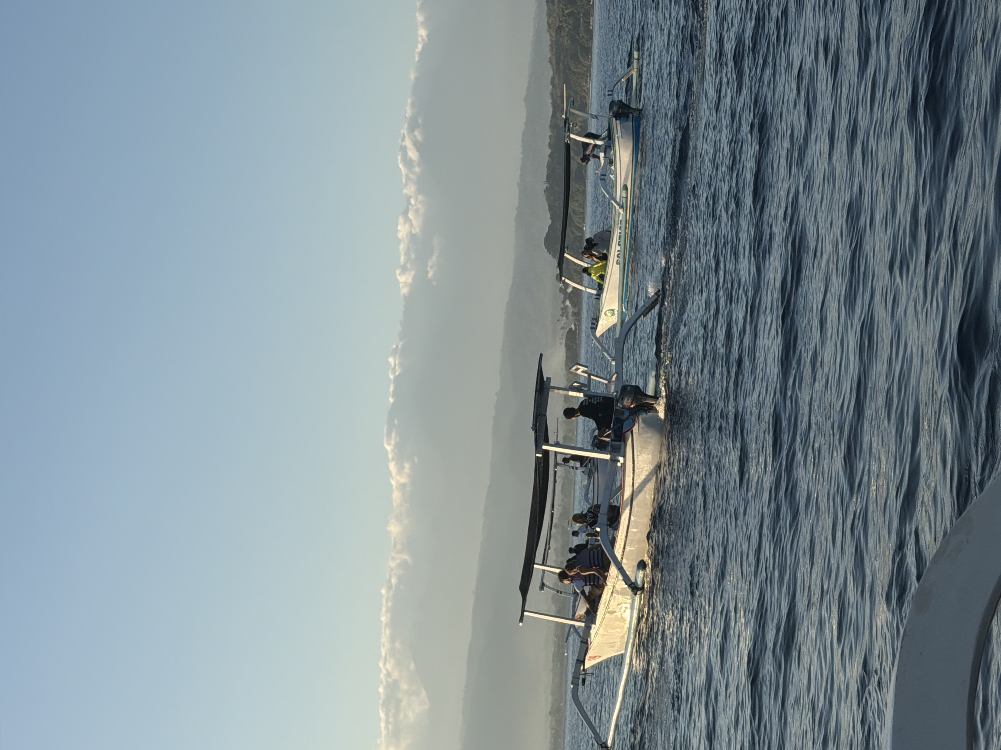 In Seririt, Indonesia, two traditional outrigger boats, known as "jukung," navigate calm, deep blue waters under a clear, pale blue sky with scattered white clouds forming a hazy line above distant, subdued green mountains. Both boats are white with a dark canopy supported by a wooden frame, and feature long, stabilizing outriggers on either side. Several individuals are seated within each boat. The boat in the foreground, on the left, has its number "29" visible on its hull. A person is seated at the rear of this boat, appearing to steer. The second boat, to the right and slightly further back, also carries passengers, one of whom is leaning over the side, possibly engaged with the water. The sun appears to be low in the sky, casting a gentle light and highlighting the ripples on the water's surface. The overall scene depicts a peaceful maritime activity near the coast.