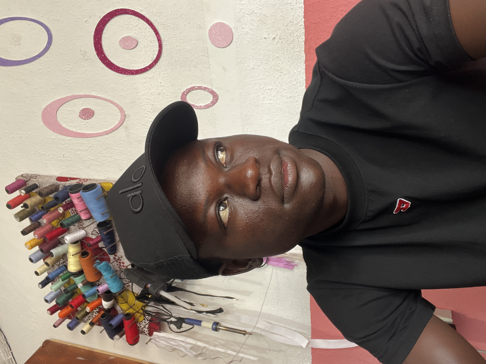 A man wearing a black baseball cap with the letters "ALO" and a black t-shirt with a red and white "P" emblem is in a room in Badagry, Nigeria. Behind him, a wall is decorated with colorful circular patterns. Spools of thread are attached to the wall as well. Various objects, including wires, a purple zipper, and a soldering iron, are on a white surface beneath the spools of thread. The wall is primarily white, but includes a vertical red stripe.