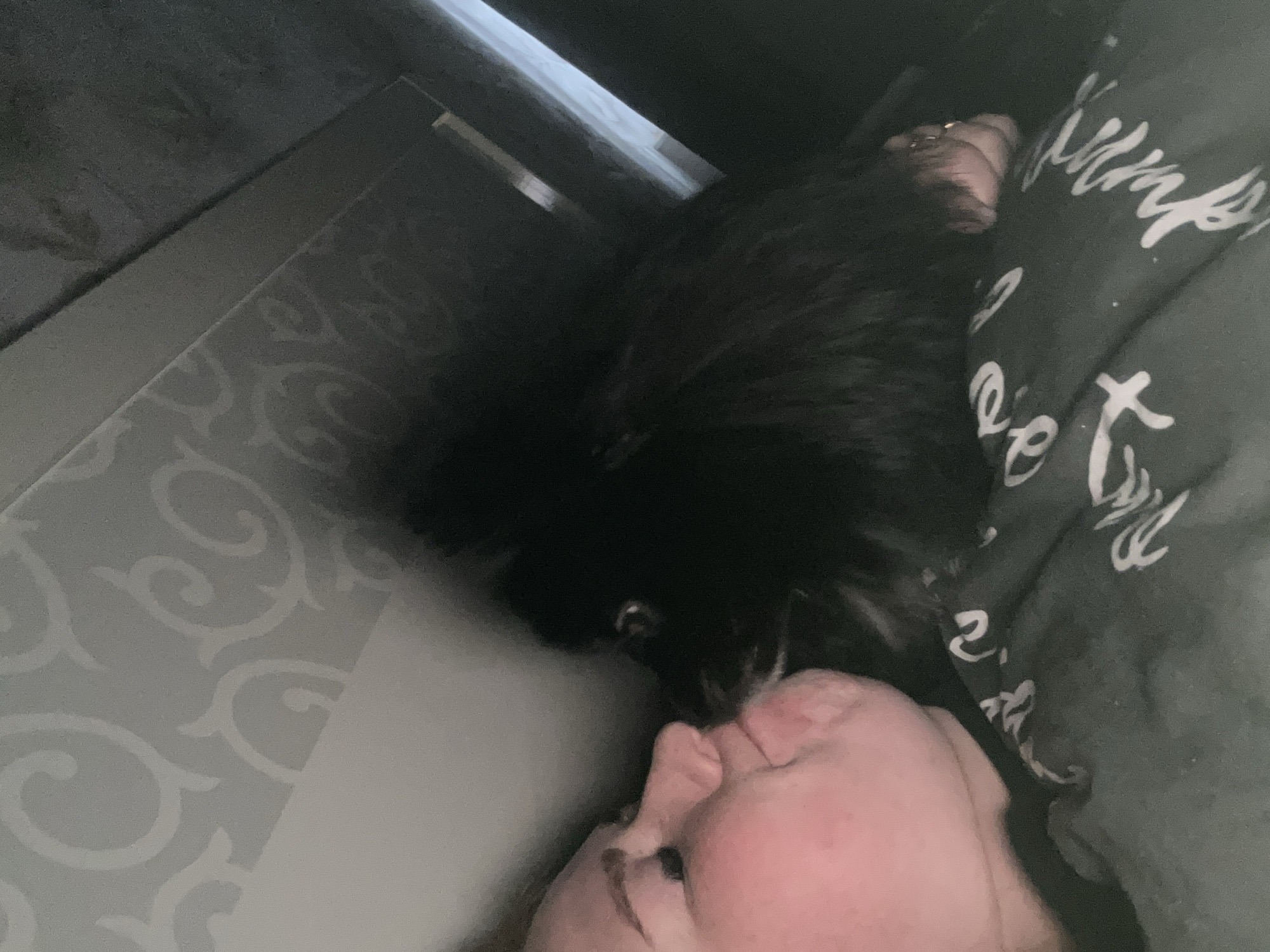 A close-up shot depicts a person's face with their eyes closed, lying next to a fluffy black dog. The person is on a patterned gray and white surface, likely a bed. Part of a gray blanket with white writing is visible beside the person. The photo was taken indoors. The location is Chaikovsky, Russia.