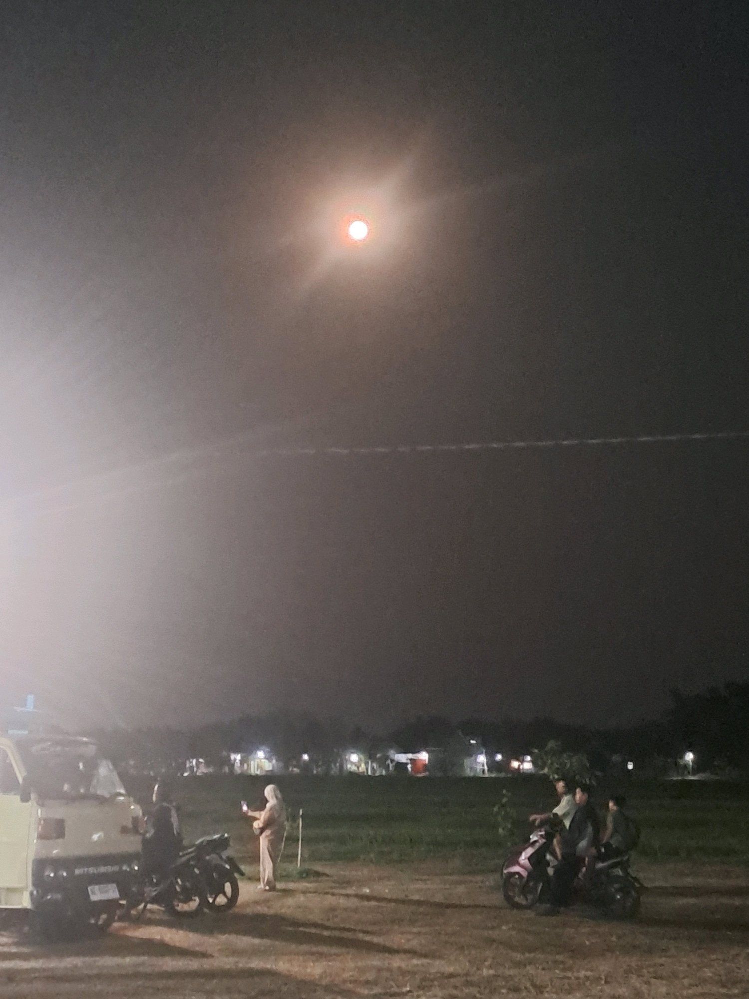 The image depicts a nighttime scene in Sumberejo, Indonesia, under a hazy, reddish-orange full moon that is positioned in the upper center of the frame, emitting a soft glow. In the foreground, a light-colored van, possibly a pick-up truck, is parked on the left. To the right of the van, a person wearing a light-colored headscarf is standing and taking a photograph with a mobile phone. Next to the van, a dark-colored motorcycle is parked. Further to the right, a pink and black motorcycle carries three individuals, with the rider in the front and two passengers behind. The background consists of a dimly lit grassy field with scattered trees and distant structures, possibly buildings with illuminated windows. The sky exhibits a thin, horizontal trail, suggestive of an airplane or contrail. The overall atmosphere is serene and suggests a gathering or casual outing in an open area at night.