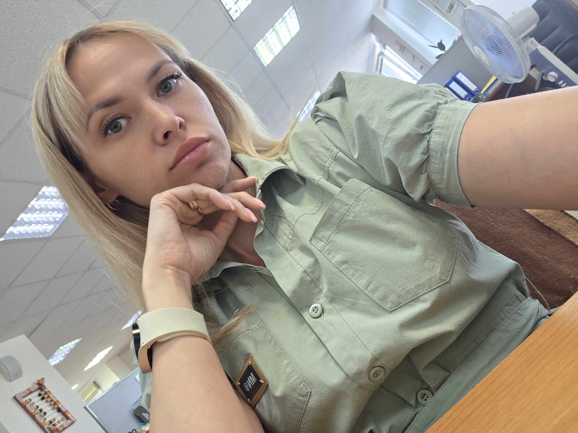 A blonde woman is taking a selfie. She is wearing a light green button-up shirt with a patch on the pocket that says "WM" in Cyrillic. She is resting her chin on her hand and wearing a white smartwatch. The photo is taken from a low angle. The background shows an office setting with fluorescent lighting, a window, and a fan. The location is likely an office in Chaikovsky, Russia, inferred from the Cyrillic patch.