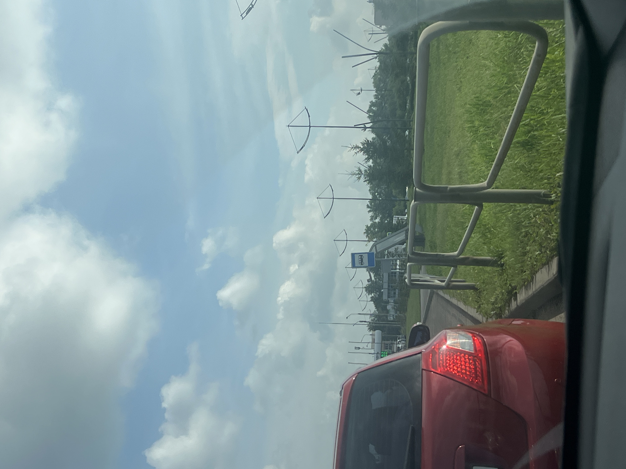 The image captures a view from inside a vehicle in Magnitogorsk, Russia, looking towards a roadside scene on a partly cloudy day. In the foreground, the rear of a red hatchback is visible, with its taillights illuminated. To the right, a metal guardrail lines a grassy verge. Beyond the guardrail, a row of stylized, abstract sculptures resembling elongated, curved triangles on poles stand against a backdrop of green trees and buildings. A blue sign with a white pictogram of a bus indicates a bus stop. Further in the distance, a gas station with visible pumps and signage is present. The sky is filled with scattered white clouds against a light blue expanse.