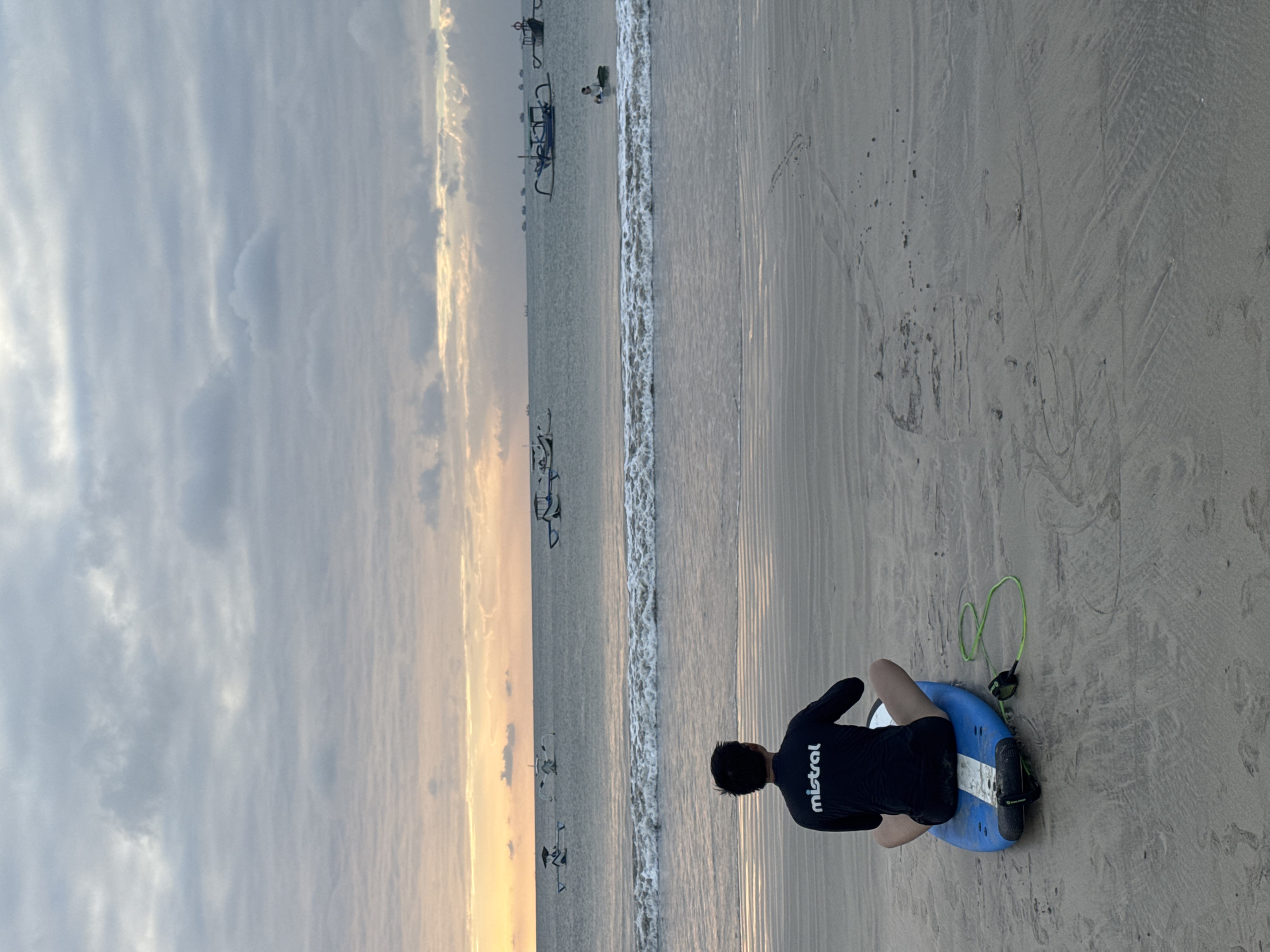 A person sits on a blue and white surfboard on a sandy beach in Kuta Selatan, Indonesia, facing the ocean. The person is wearing a black long-sleeved rashguard with "mistral" printed in white on the back. The surfboard has a green leash attached. The ocean has moderate waves and several traditional Indonesian fishing boats are visible on the horizon. The sky is overcast with soft, diffused light indicating either dawn or dusk. The sand on the beach shows ripples and some debris.