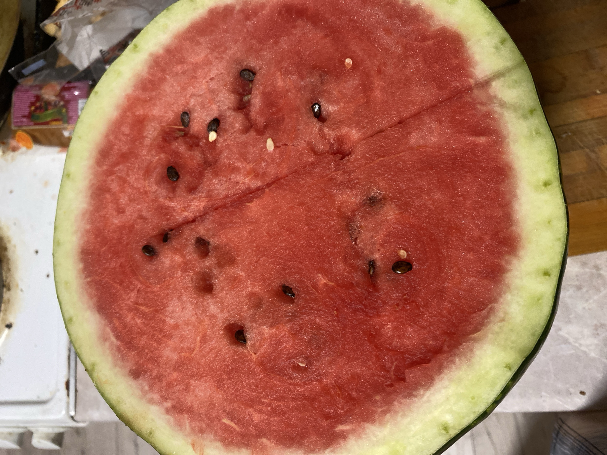 A close-up, overhead shot shows a cut watermelon. The flesh is a vibrant red, with scattered black seeds and a few lighter, immature seeds. The rind is light green with a slightly darker green outer edge. The watermelon is positioned on a wooden surface, with a white appliance and a colorful package visible in the blurred background. The scene is indoors, suggesting a domestic setting. There are no people present in the image. The location within the city of Chornomorsk, Ukraine, is not discernible from the provided image.