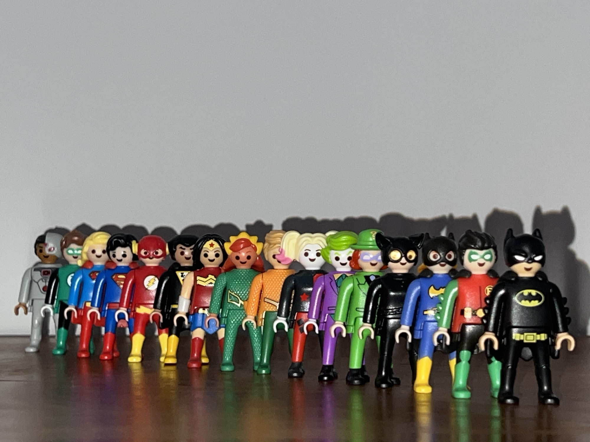 A collection of 14 Playmobil figures of DC Comics superheroes and villains are arranged in a line on a polished, dark wooden surface against a light gray background. From left to right, the figures are: Cyborg, Green Lantern, Superman, Flash, Batman, Wonder Woman, Aquaman, Bumblebee, Harley Quinn, the Joker, the Riddler, Catwoman, Batgirl, and Batman. The figures are standing upright and facing forward. The lighting is bright and even, casting shadows of the figures on the background. The location is in the city of Unknown, Unknown.