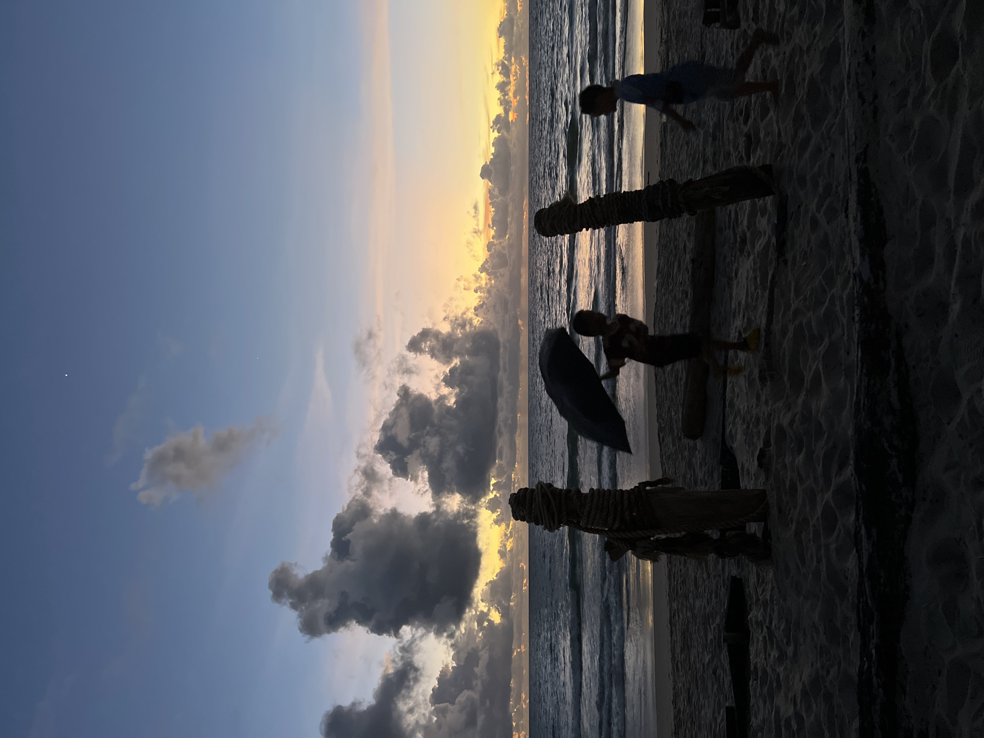 This is a vertical, wide-angle, eye-level shot of a beach scene at sunset. The sun is low on the horizon, casting a warm, golden glow across the sky, which is transitioning from a soft blue to vibrant oranges and yellows. Dark, textured clouds are scattered across the sky, creating a dramatic contrast with the bright sunset. The ocean is calm, with gentle waves rolling onto the sandy shore.
In the foreground, silhouetted figures of people are visible. Two children appear to be playing on the beach. One child, wearing a blue shirt, is running or jumping in the sand. Another child is holding a dark umbrella. Two wooden posts, wrapped in rope, stand vertically on the beach, adding a rustic element to the scene. The sand is finely textured, with footprints and ripples visible.
The scene evokes a sense of peace and tranquility, with the beautiful sunset and the playful activity of the children creating a harmonious atmosphere. The light suggests it is late afternoon or early evening. The location appears to be a tropical beach due to the palm-like posts and the warm, sandy environment. There is no visible text in the image.