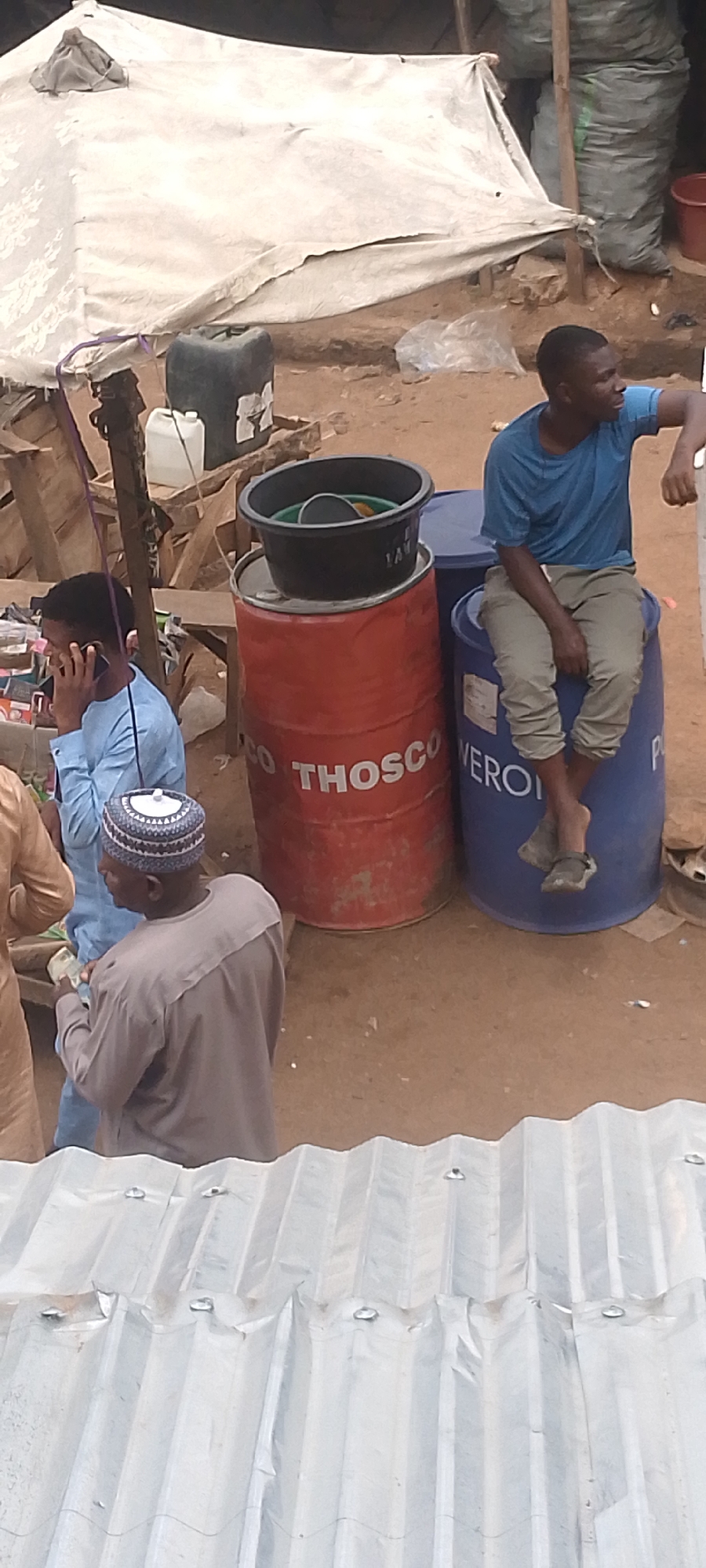 In Bukuru, Nigeria, a daytime outdoor scene shows several people and objects. A man in a blue shirt and beige pants is seated on a blue barrel marked 'VEROI'. His right arm is resting on his knee. Next to the blue barrel is a red barrel with the word 'THOSCO' written on it. A black bucket sits on top of the red barrel. A man in a light blue outfit speaks on a cell phone near the barrels. Another man in a grey outfit with a white cap is standing nearby, possibly observing. The surroundings suggest a market or commercial area. A white tent-like structure is visible behind the barrels, along with what appears to be bags of goods. Corrugated metal sheets are present in the foreground.