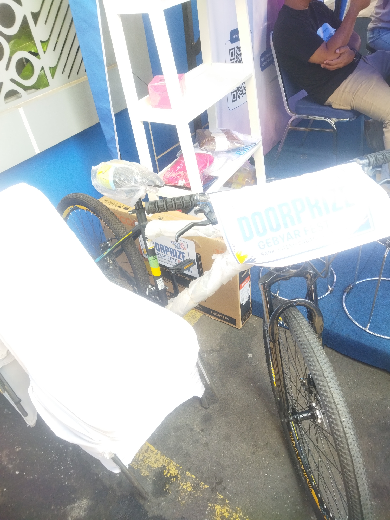 The image shows a close-up view of a bicycle with a "DOORPRIZE GEBYAR FEST BANK JATENG" sign attached to its handlebars. The bicycle is partially covered with white plastic sheeting. In the background, a person wearing a black t-shirt and tan pants is seated on a blue chair. Behind the person, a white shelf unit is visible with various items, including a pink box. The scene appears to be an outdoor event or exhibition. The presence of the "BANK JATENG" sign suggests the location might be in Giripurwo, Indonesia, as Bank Jateng is a regional bank operating in Central Java, Indonesia.