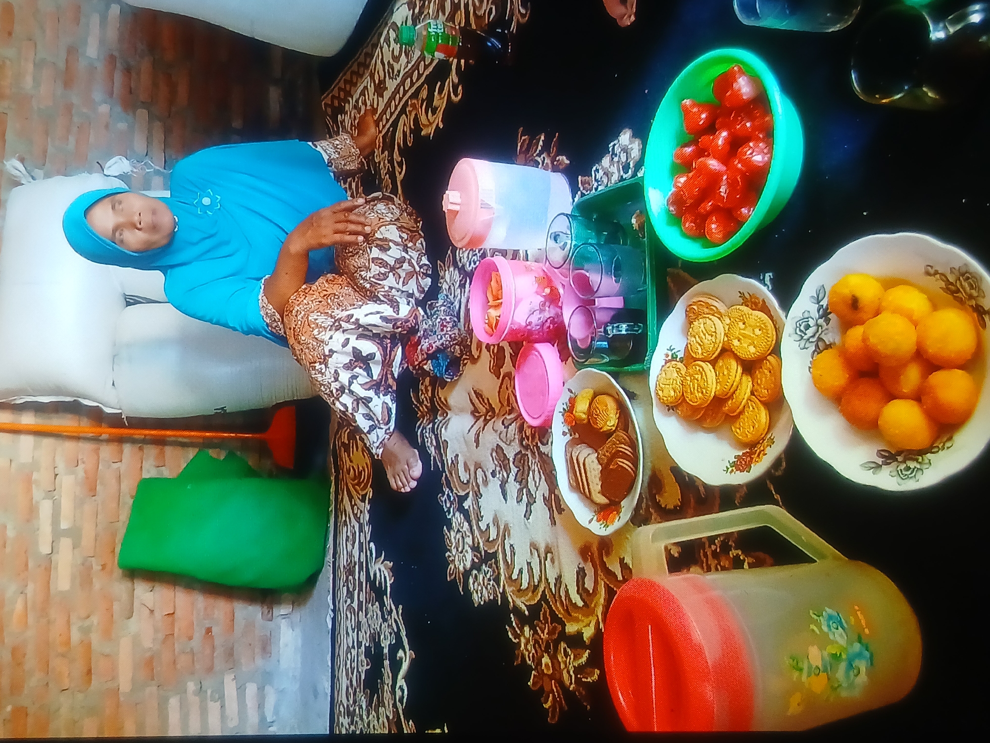The image depicts an indoor scene set on a patterned carpet. A woman wearing a blue hijab and a patterned garment is seated on the floor, looking to her right. In front of her, a variety of food items are arranged on plates and in bowls. These include cookies, what appear to be small pastries or fruits, and red, heart-shaped candies. There are also several plastic containers and cups. To the left of the woman, a large green fabric object is propped against a brick wall, next to a broom. In the background, large white sacks are visible, and the wall is constructed of exposed bricks. A bottle of soda is placed on the carpet. The overall impression is of a domestic setting where food is being served or shared. The location is specified as Setia Bakti, Indonesia.