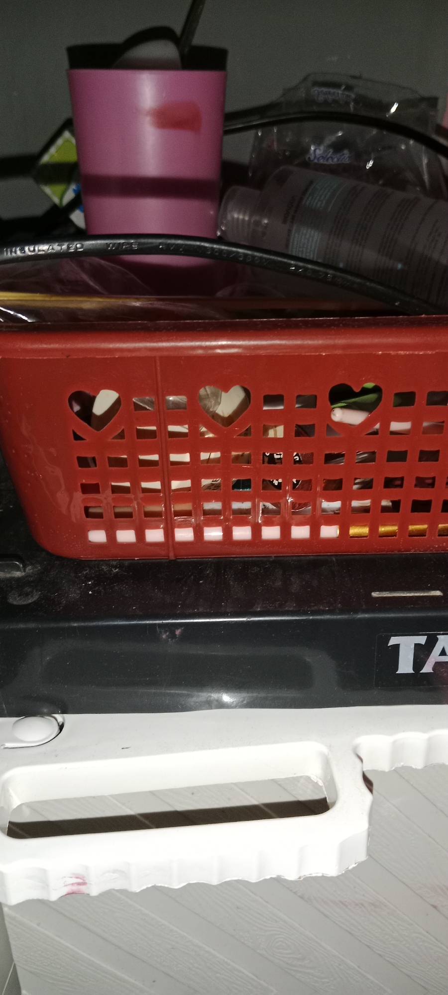 The image shows a close-up, eye-level view of a red plastic basket with heart-shaped cutouts on its sides, positioned on top of a black appliance. The basket contains various small items that are partially visible through the cutouts and the grid pattern of the basket. Behind the basket, a pink cup is visible, along with other blurred objects, possibly on a shelf. The black appliance has a visible logo that partially reads "TA". The foreground shows a white object with a textured surface, possibly a piece of furniture or another appliance, with a cutout handle. The overall lighting is dim, suggesting an indoor setting. There are no people visible in the image. The scene appears to be a domestic or commercial storage area. The location in Subang, Indonesia, is not determinable from the visual information provided.
