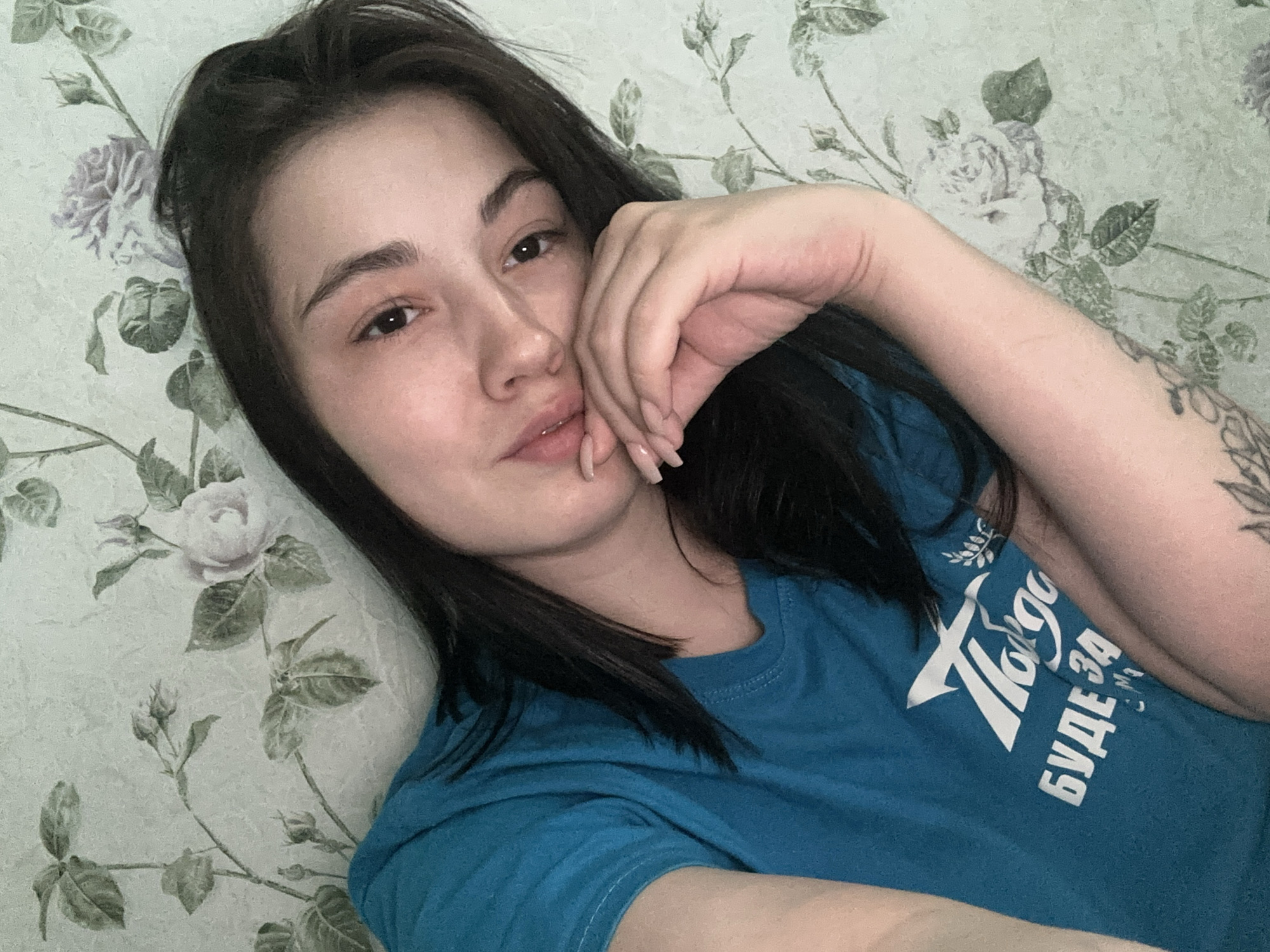 A young woman is lying down, propped up, likely on a bed or sofa covered in a floral patterned material that appears to be stylized roses and leaves. She is resting her hand against her face. She is wearing a blue t-shirt with white text, possibly in Cyrillic. A tattoo is visible on her upper left arm. The location is Chaikovsky, Russia.